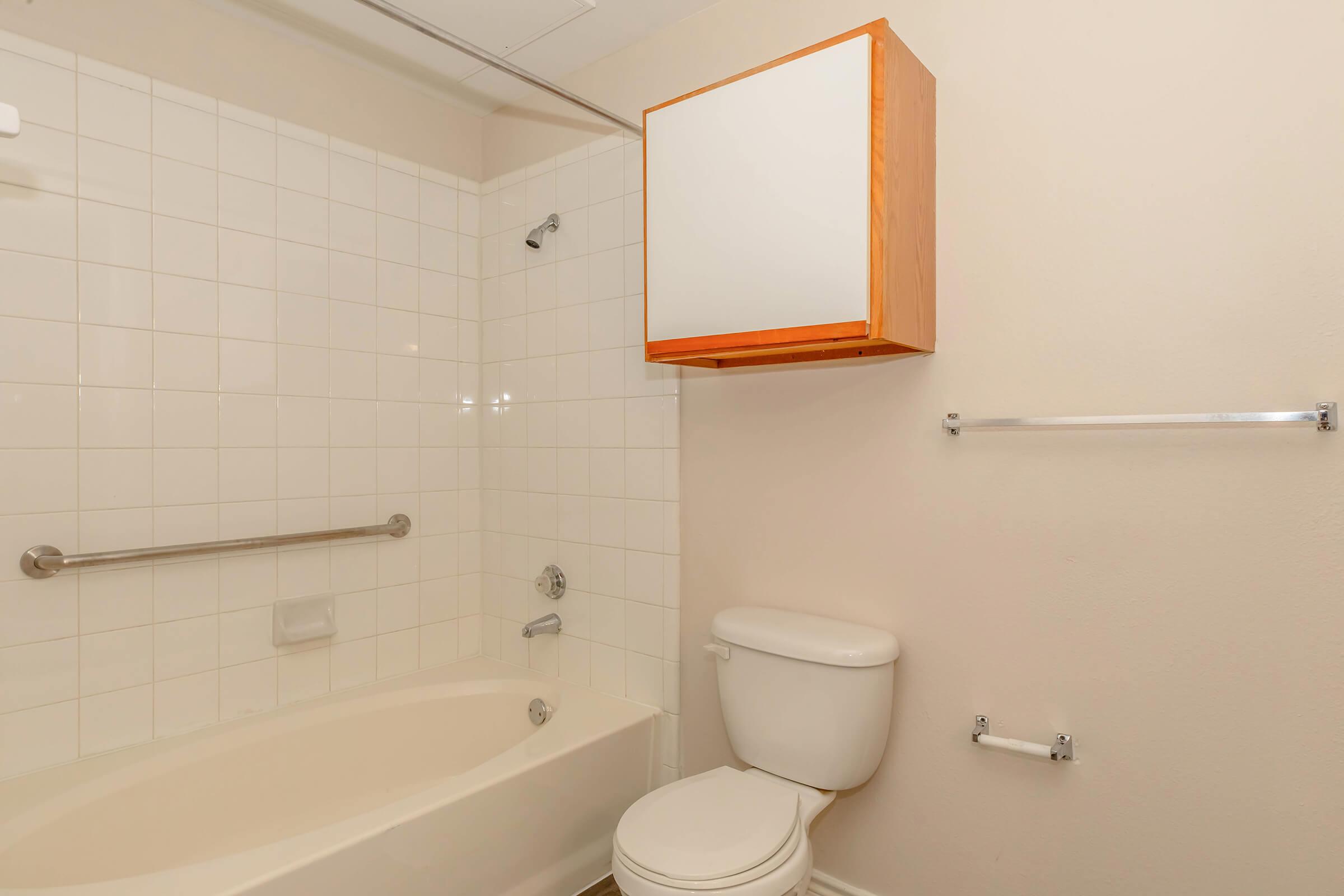A clean and simple bathroom featuring a bathtub with a showerhead, a toilet, and a wooden cabinet mounted on the wall. The walls are tiled in white, and there is a towel bar on one side. Natural light is soft, highlighting the minimalist design.