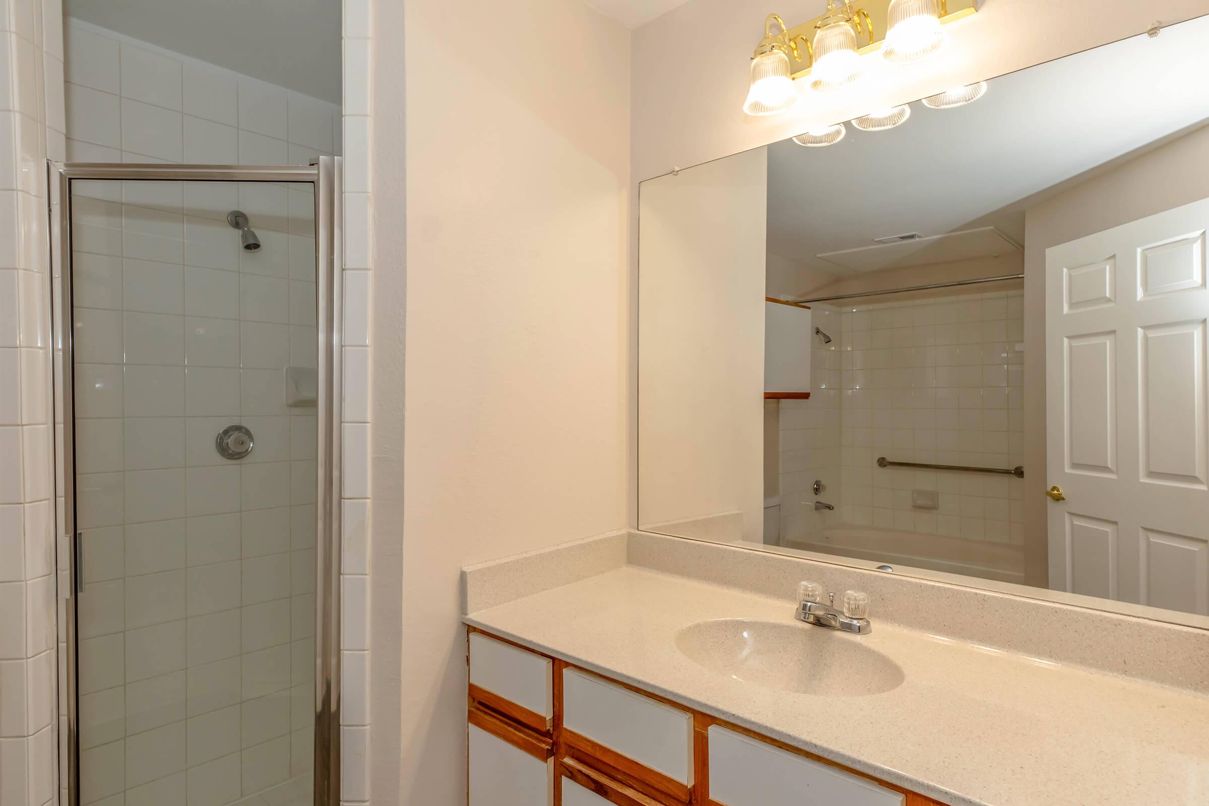 A clean, well-lit bathroom featuring a glass shower enclosure, a white countertop sink with a single faucet, and a large mirror above the sink. The bathroom has light-colored walls and a door leading to another area. There are towel bars and ample space, creating a functional and tidy appearance.