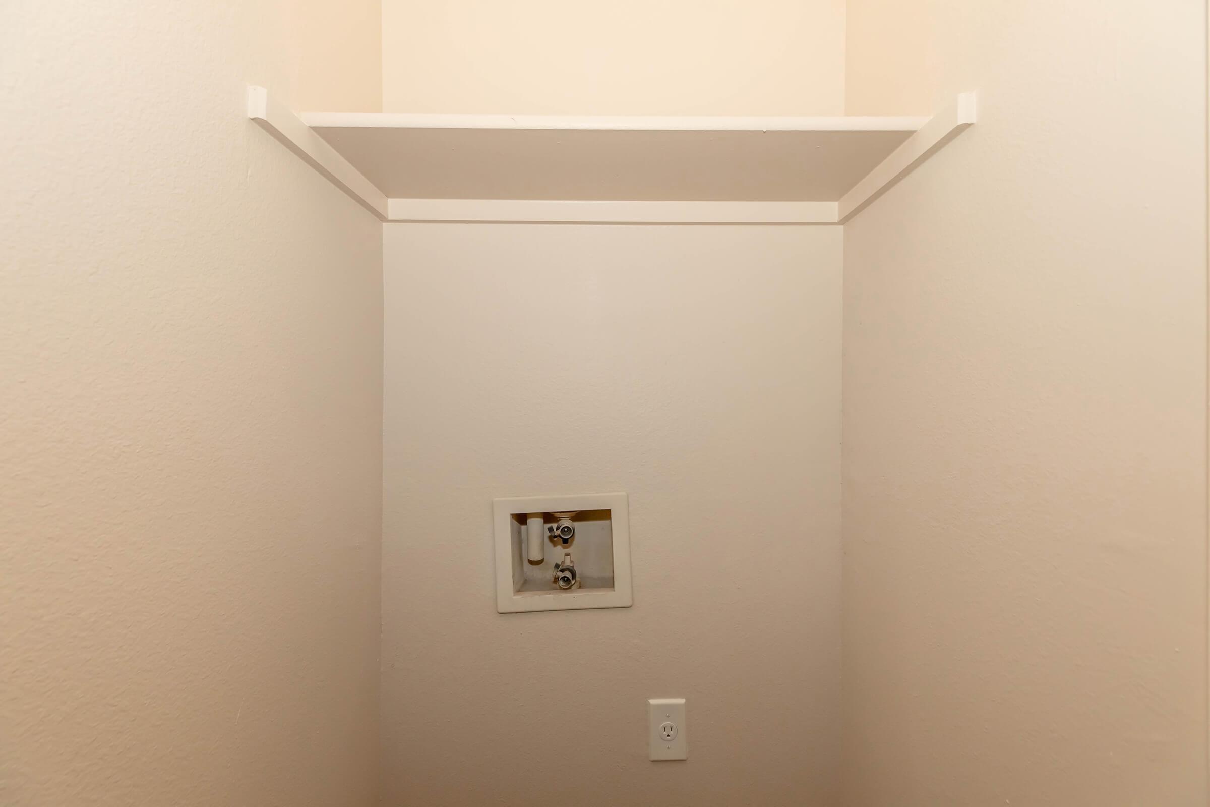 A small, empty closet with plain walls painted in a light color. A shelf is mounted at the top, and there is a small, white panel on one wall, likely for plumbing access. An electrical outlet is situated lower on the wall. The overall space is clean and uncluttered.
