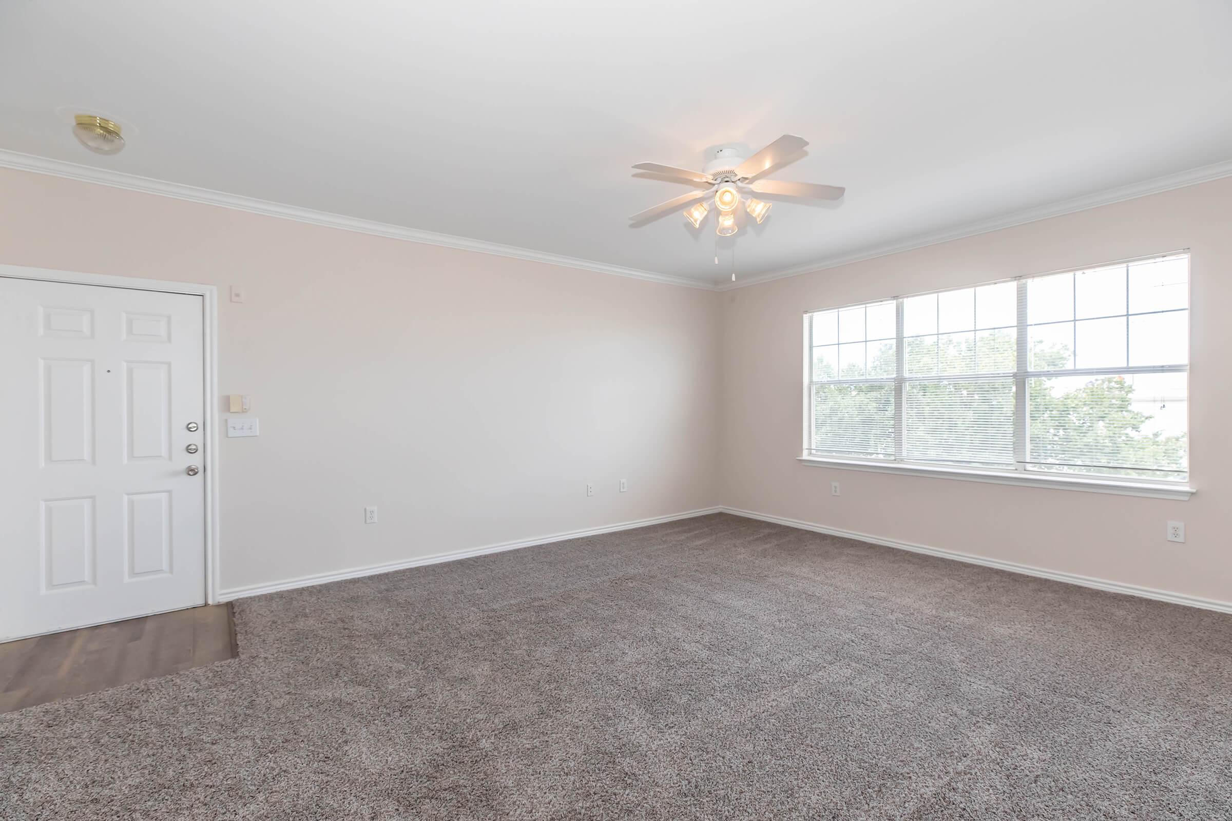 A spacious, empty room with light-colored walls and carpeted flooring. There's a ceiling fan with light fixtures, a large window allowing natural light, and a closed door leading outside. The room has a minimalist design, suitable for various furniture arrangements.