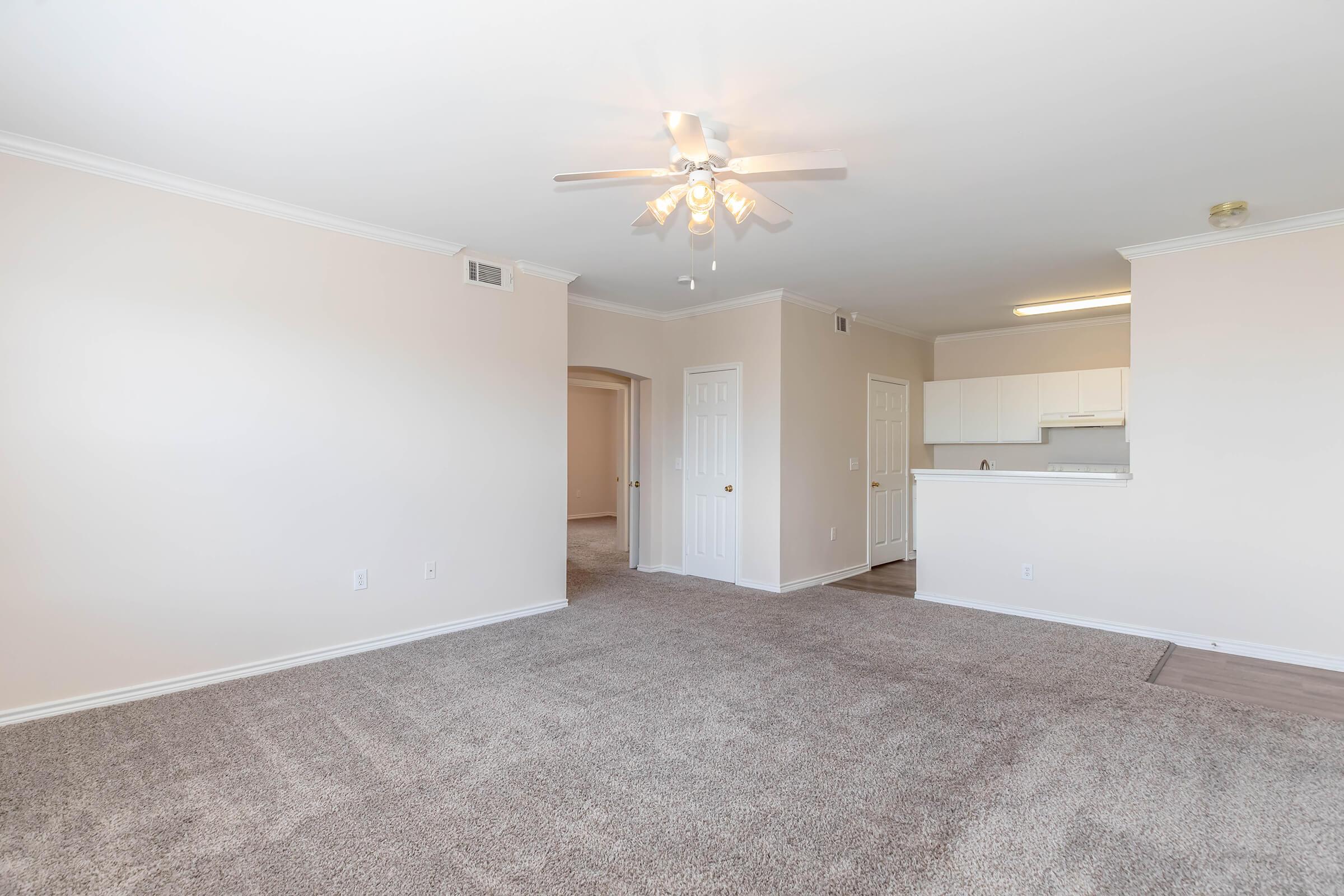 A spacious, carpeted living room with neutral-colored walls and a ceiling fan. An archway leads to another room, while a kitchen area with white cabinetry is visible in the background. Natural light brightens the space, creating a warm and inviting atmosphere.