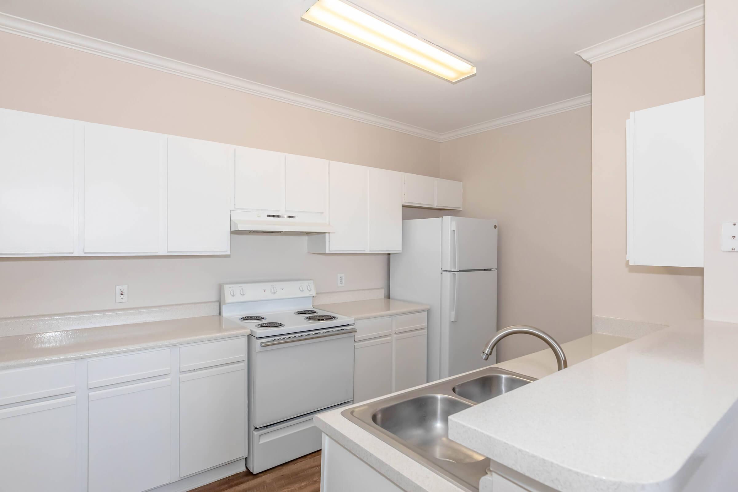 A modern kitchen featuring white cabinetry, a stainless steel sink, and a refrigerator. The layout includes a stove and oven combination, with a countertop and ample cabinet space. The walls are painted in a light neutral color, and there is bright overhead lighting.