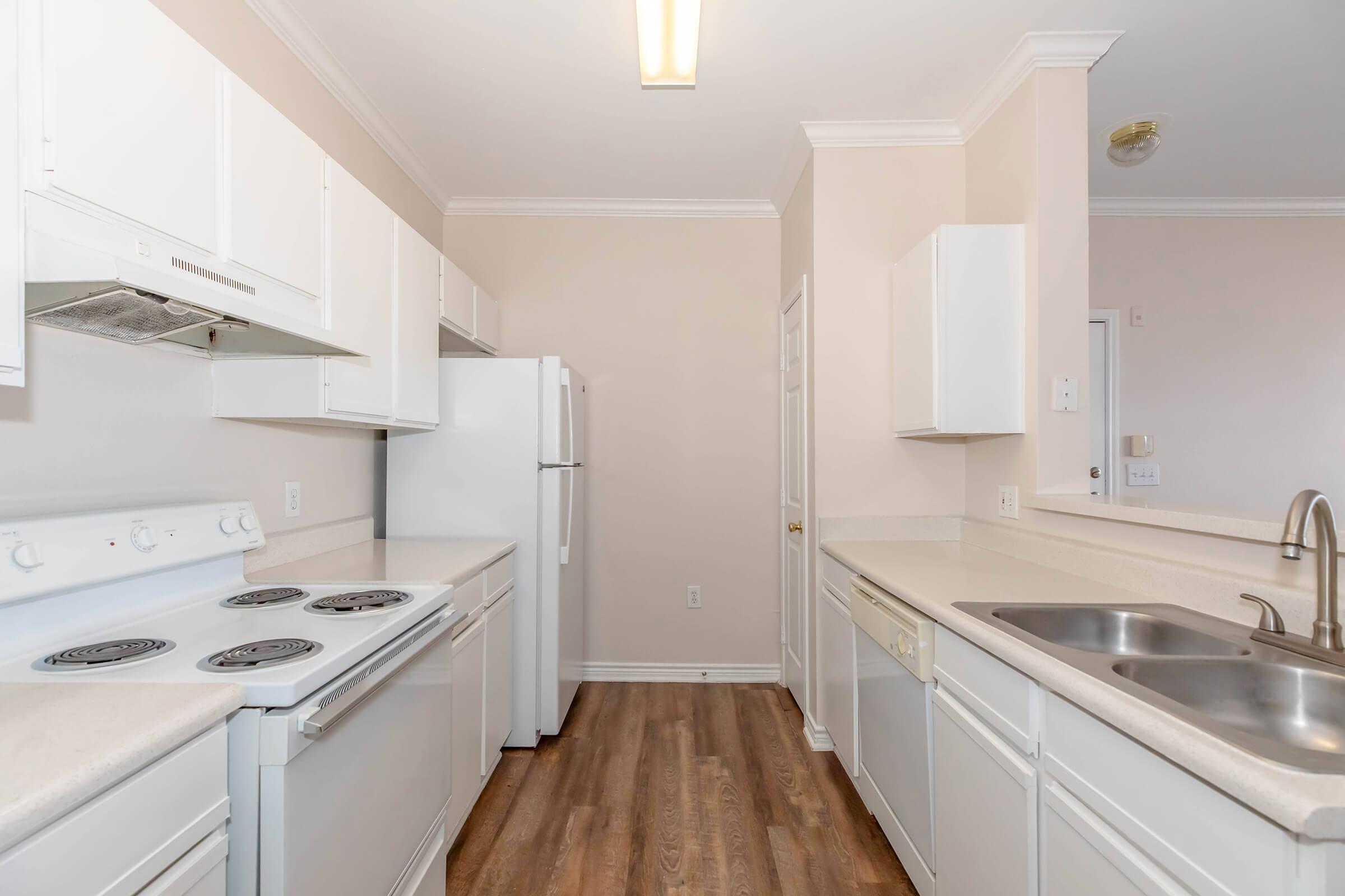 Bright, modern kitchen featuring white cabinets and appliances, including a refrigerator, stove, and dishwasher. The countertops are light-colored, and the flooring is wood-like. Natural light enters from a ceiling fixture, creating a clean and inviting atmosphere. Spacious layout with ample storage options.