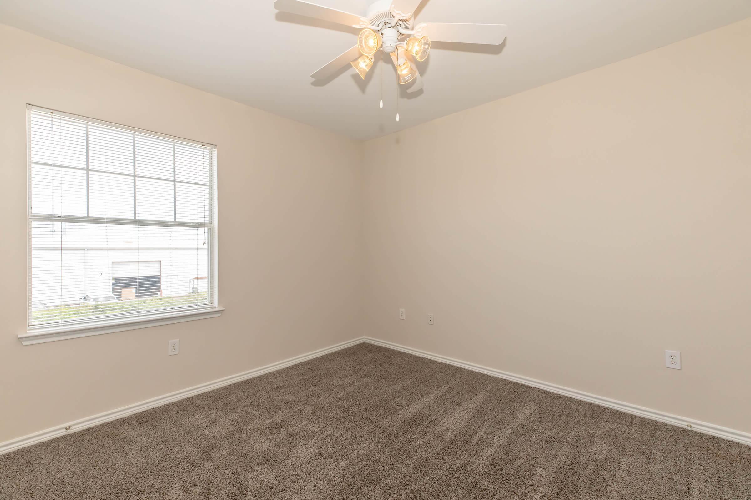 An empty room with light beige walls and a ceiling fan with four light bulbs. The floor is covered in light-colored carpet, and there is a window with blinds allowing natural light to enter. The room appears spacious and well-lit.