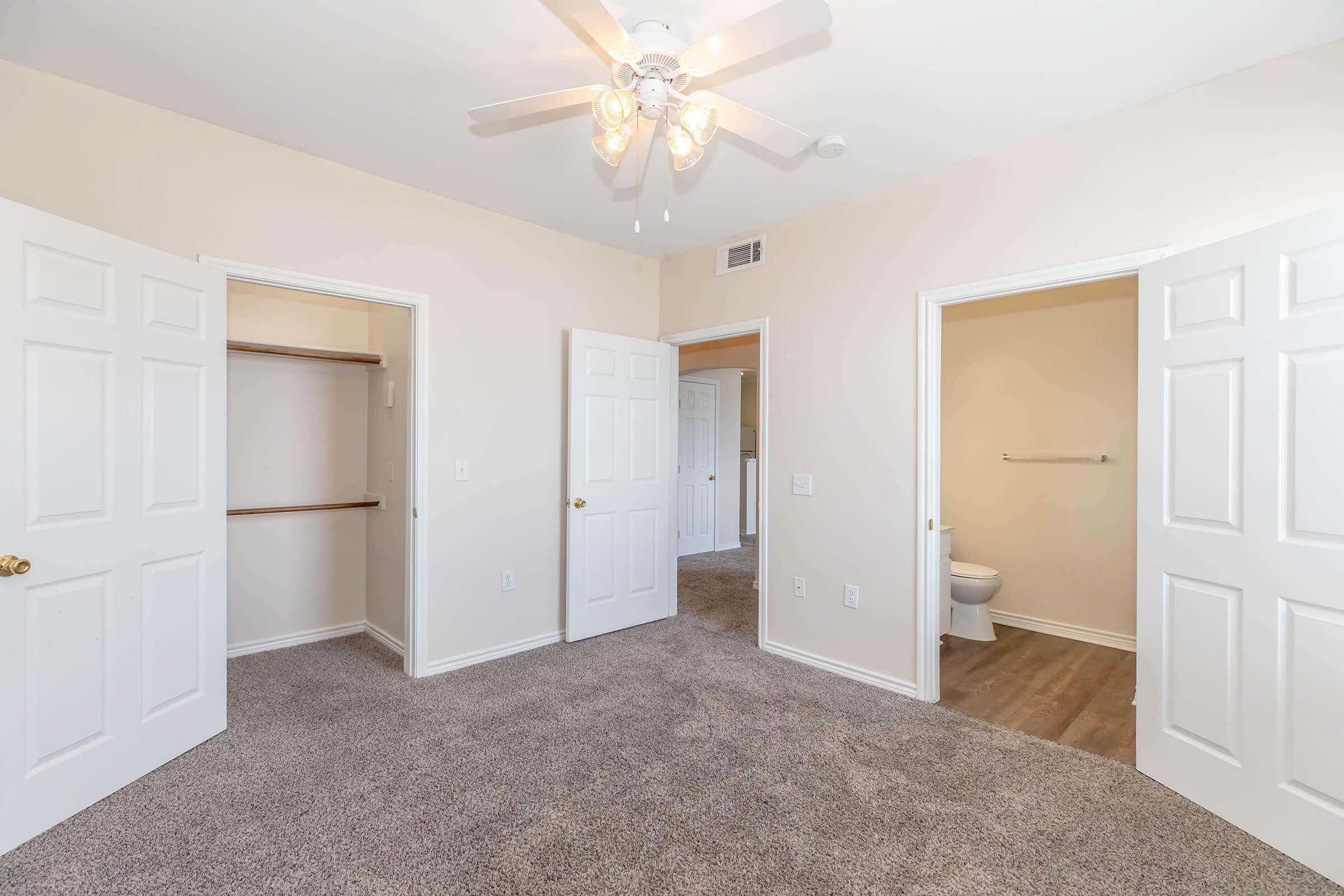 A well-lit, empty room with beige walls and carpet. Two open doors lead to closets, while another door opens to a bathroom. A ceiling fan with three lights is visible, and the overall atmosphere is clean and inviting. Perfect for a living space or bedroom.