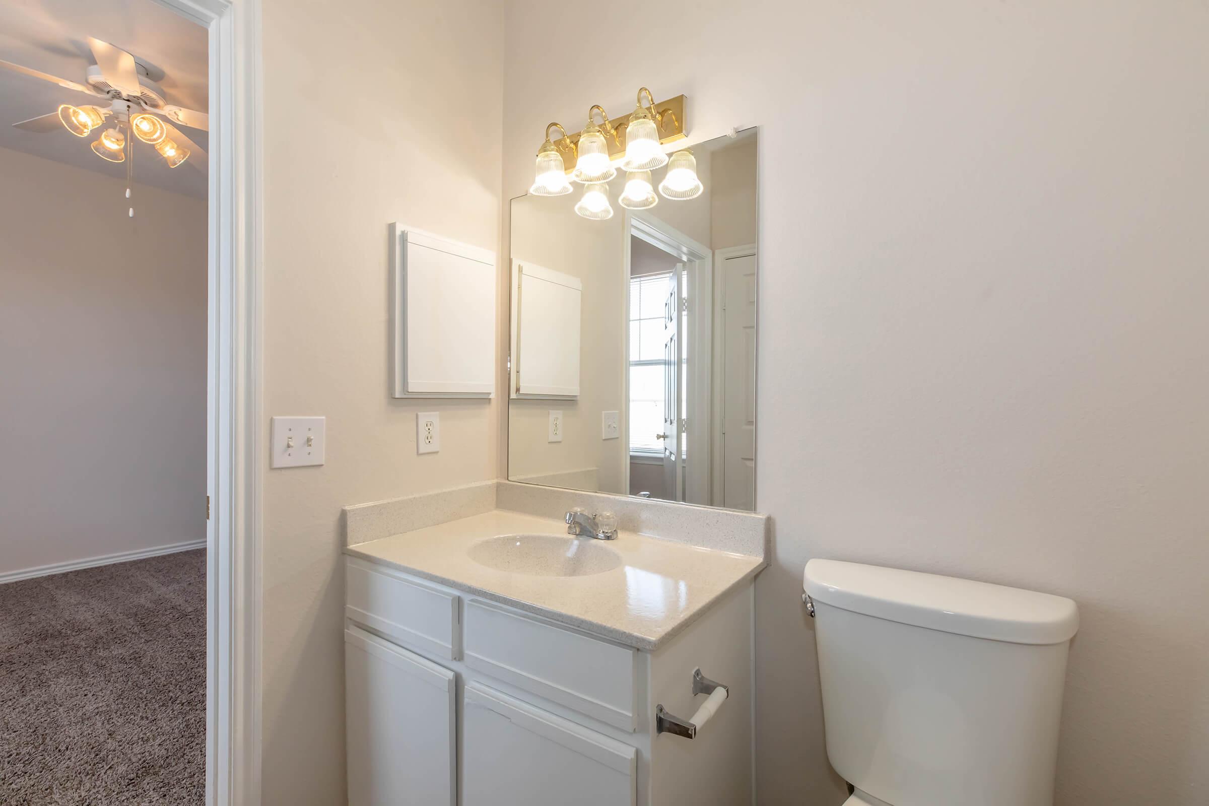 A well-lit bathroom featuring a white vanity with a sink and a mirror above it, a white toilet, and neutral-colored walls. In the background, a door leads to another room, and a ceiling fan is visible. The flooring is carpeted, providing a cozy atmosphere.