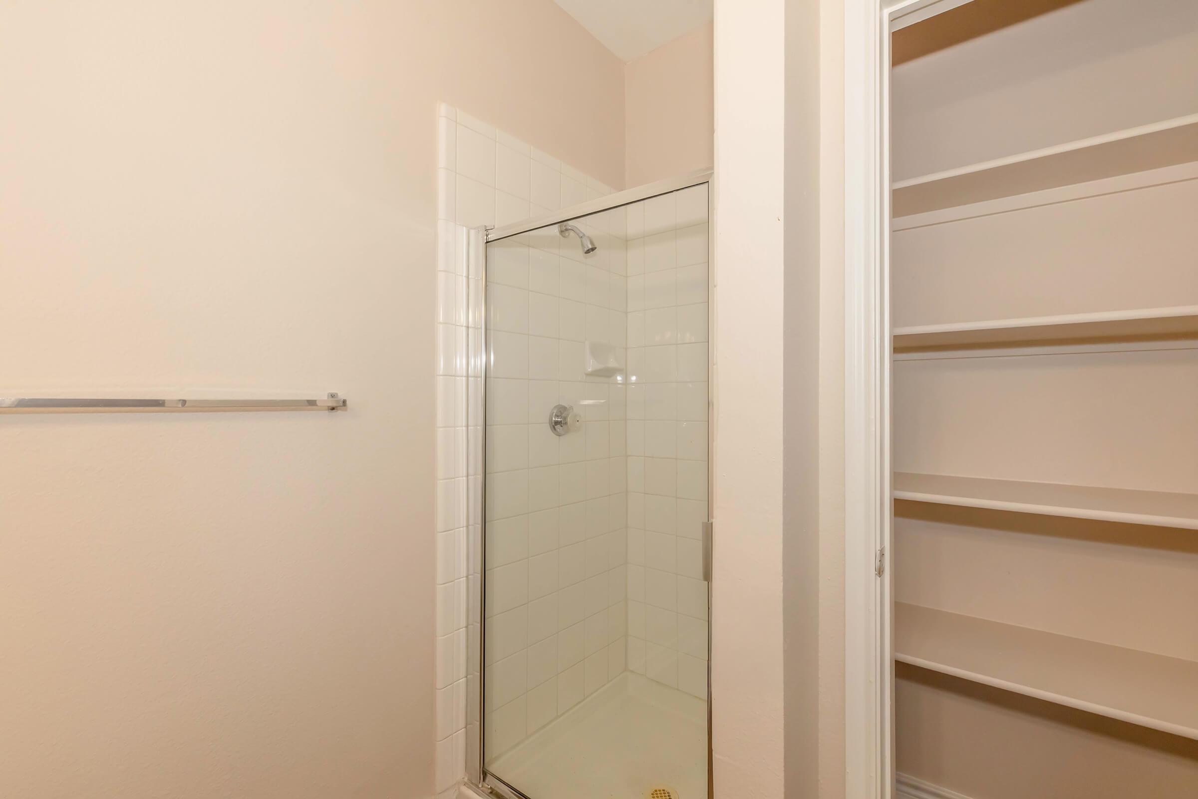 A clean and modern bathroom featuring a glass shower enclosure and a tiled floor. Light beige walls complement the space, with a white shelving unit nearby for storage. The overall design is simple and functional, suitable for any contemporary home.