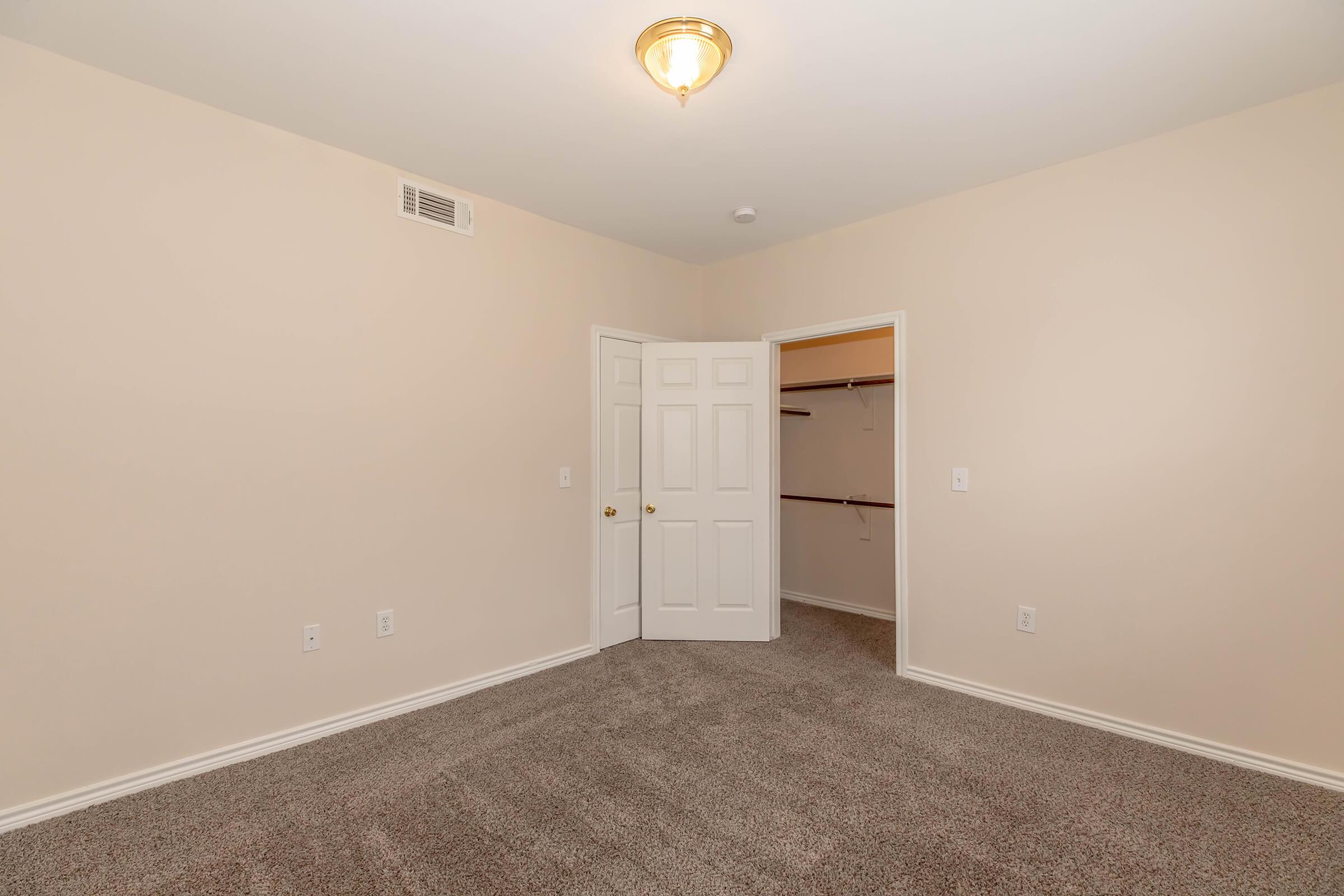 A spacious, empty bedroom featuring light beige walls, plush carpet flooring, and a single light fixture on the ceiling. There are two doors: one leading to a closet with shelving and another leading to an adjoining room. The overall ambiance is bright and neutral, suitable for various decor styles.