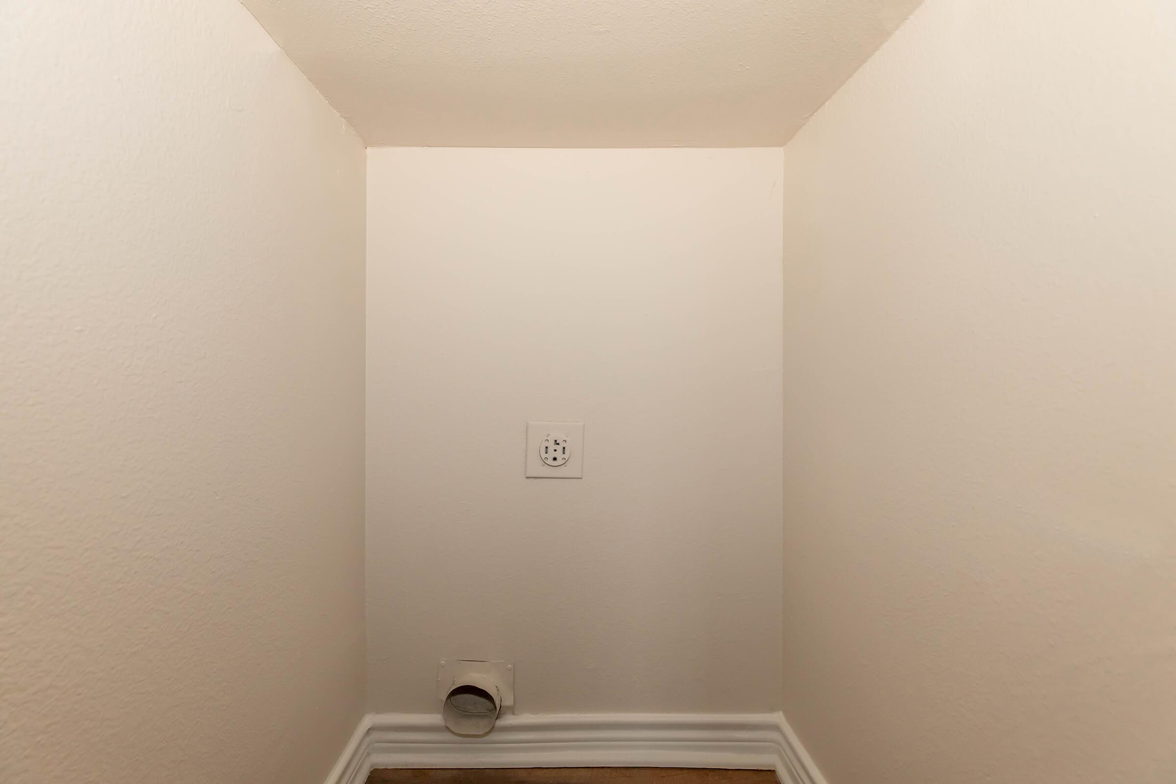A small, empty corner of a room with white walls and a wooden floor. There is an electrical outlet on the wall and a vent on the lower left side, indicating it might be a utility space or closet. The lighting is bright and the overall scene is uncluttered.