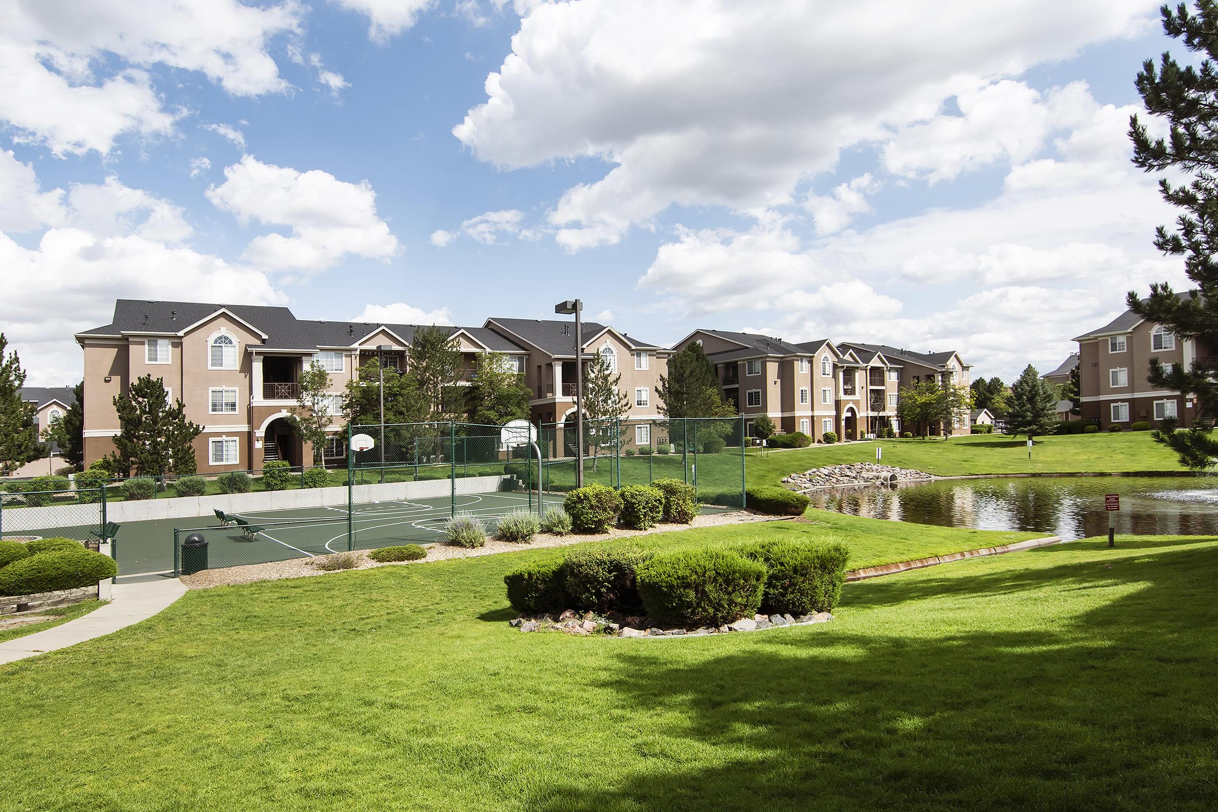A scenic view of a residential apartment complex with two-story buildings, a basketball court, and manicured lawns. In the background, there are trees and a pond under a blue sky with fluffy white clouds. The lush green landscape creates a serene outdoor environment.