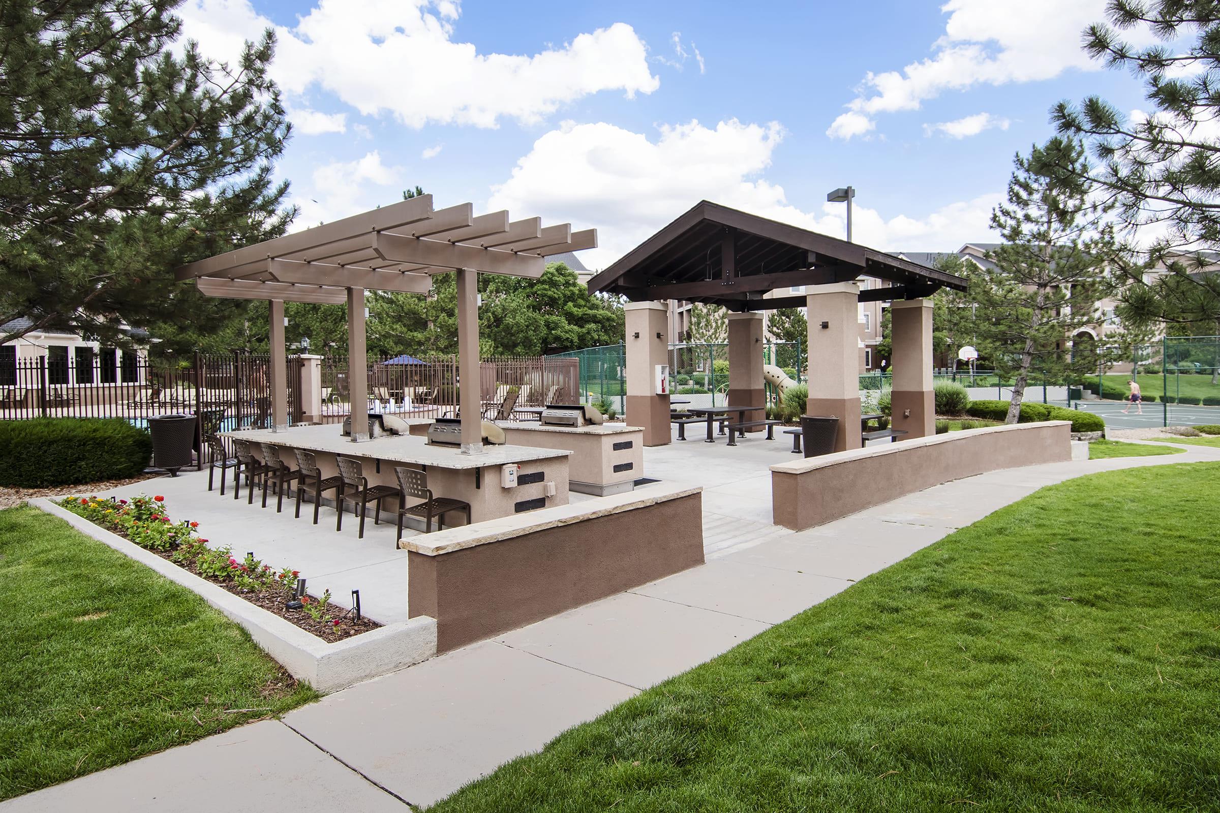Outdoor cooking and dining area featuring a covered grill station with seating, surrounded by landscaped grass and flowers. There are trees in the background and a tennis court visible in the distance. The sky is partly cloudy, creating a pleasant ambiance for outdoor gatherings.
