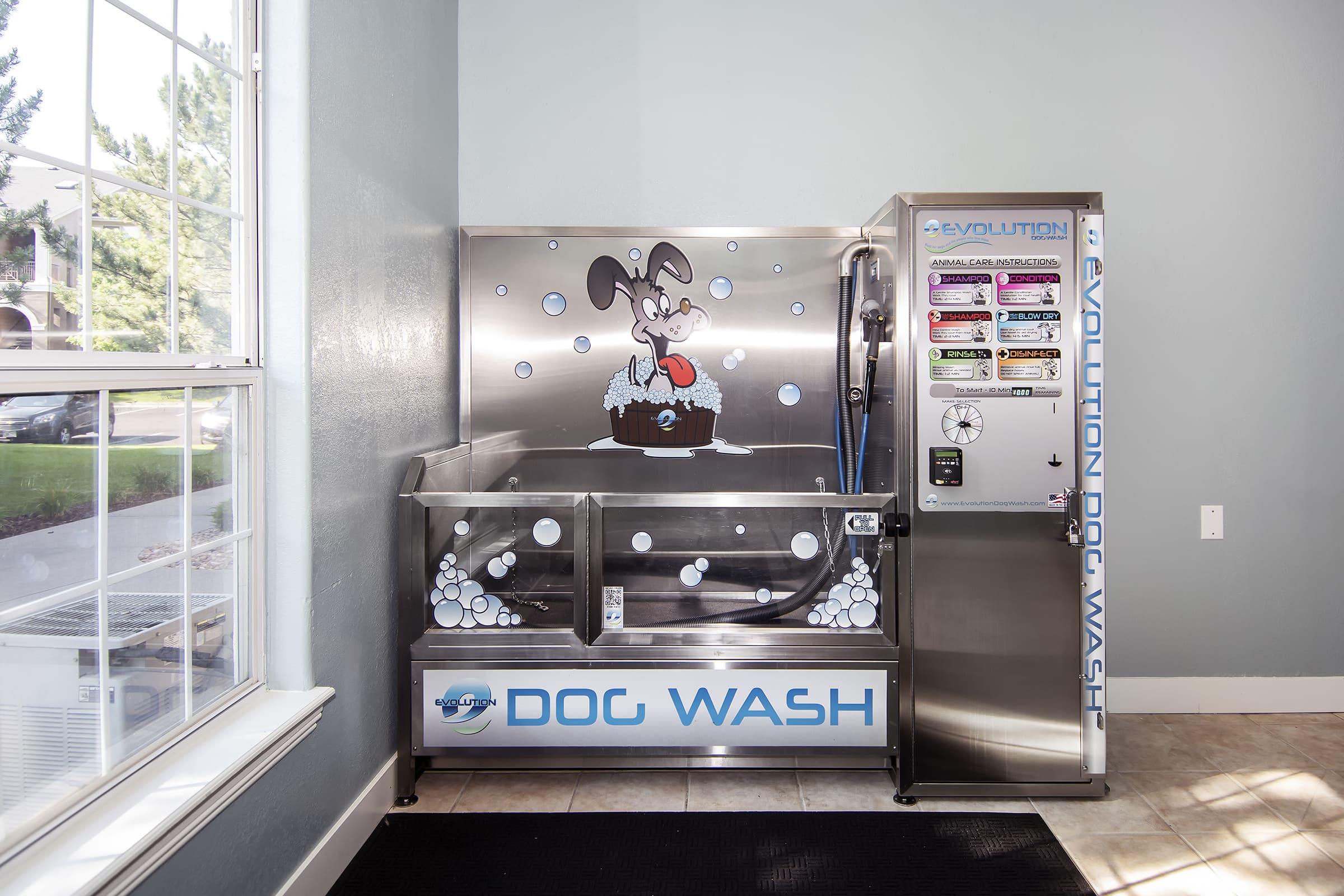 Self-serve dog wash station featuring a cartoon dog surrounded by bubbles. The stainless steel unit includes a washing area, hoses for water and shampoo, and instructions for use. Brightly lit space with window letting in natural light.