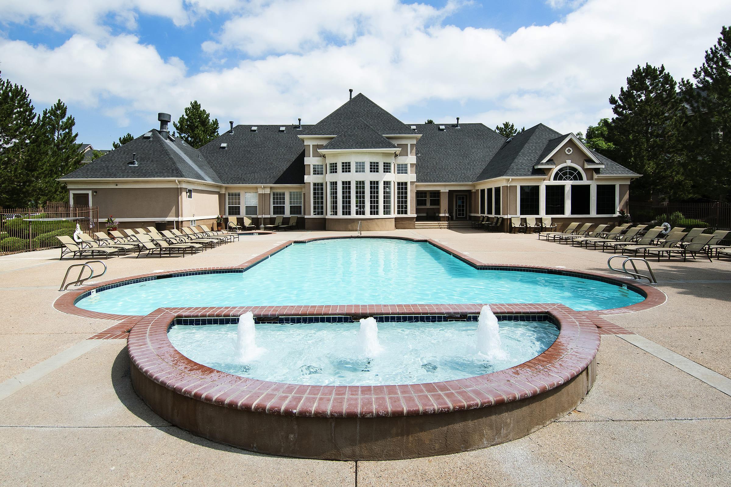A spacious outdoor swimming pool with a fountain in the foreground, surrounded by lounge chairs. In the background, a large, elegant building with multiple windows and a peaked roof is visible, set against a backdrop of trees and a partly cloudy sky.