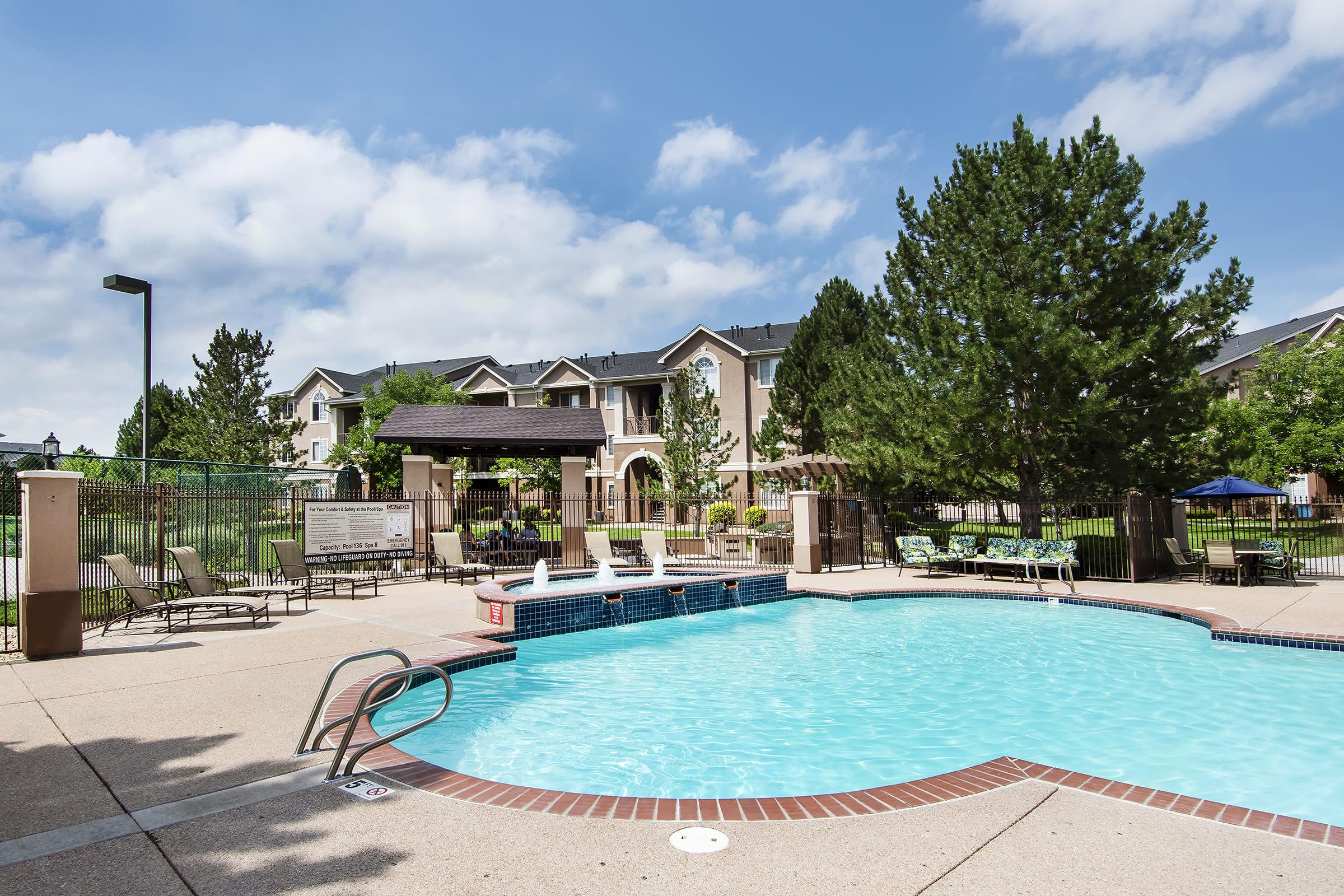 A clear blue swimming pool surrounded by lounge chairs and greenery, with apartment buildings in the background. The sky is partly cloudy, and there are umbrellas providing shade in the area. The setting appears inviting and well-maintained, perfect for relaxation.
