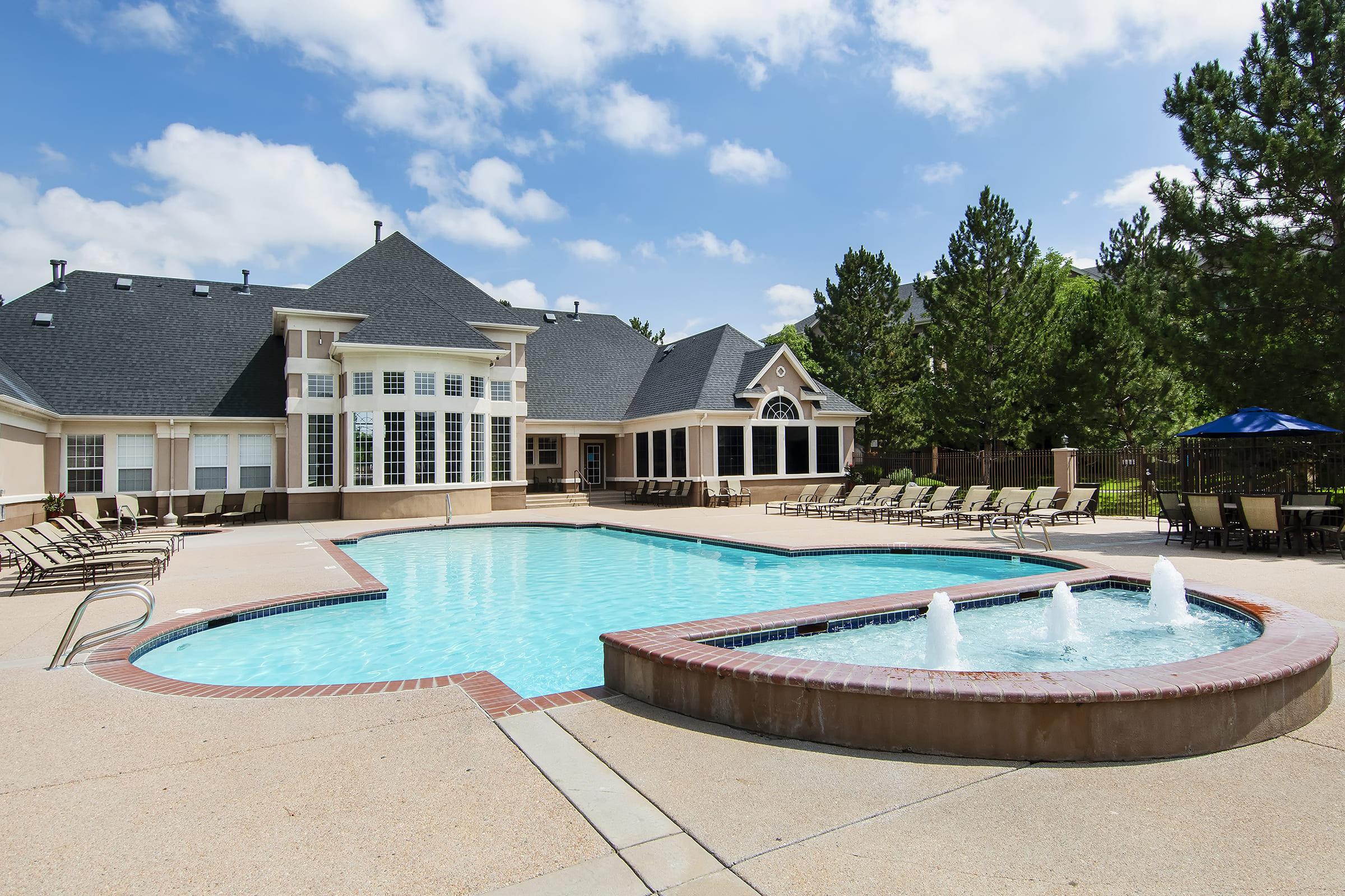 A sunny outdoor pool area featuring a large swimming pool with fountains, surrounded by lounge chairs. The backdrop includes a stylish building with multiple windows and a peaked roof, along with lush green trees lining the space. Perfect for relaxation and recreation.