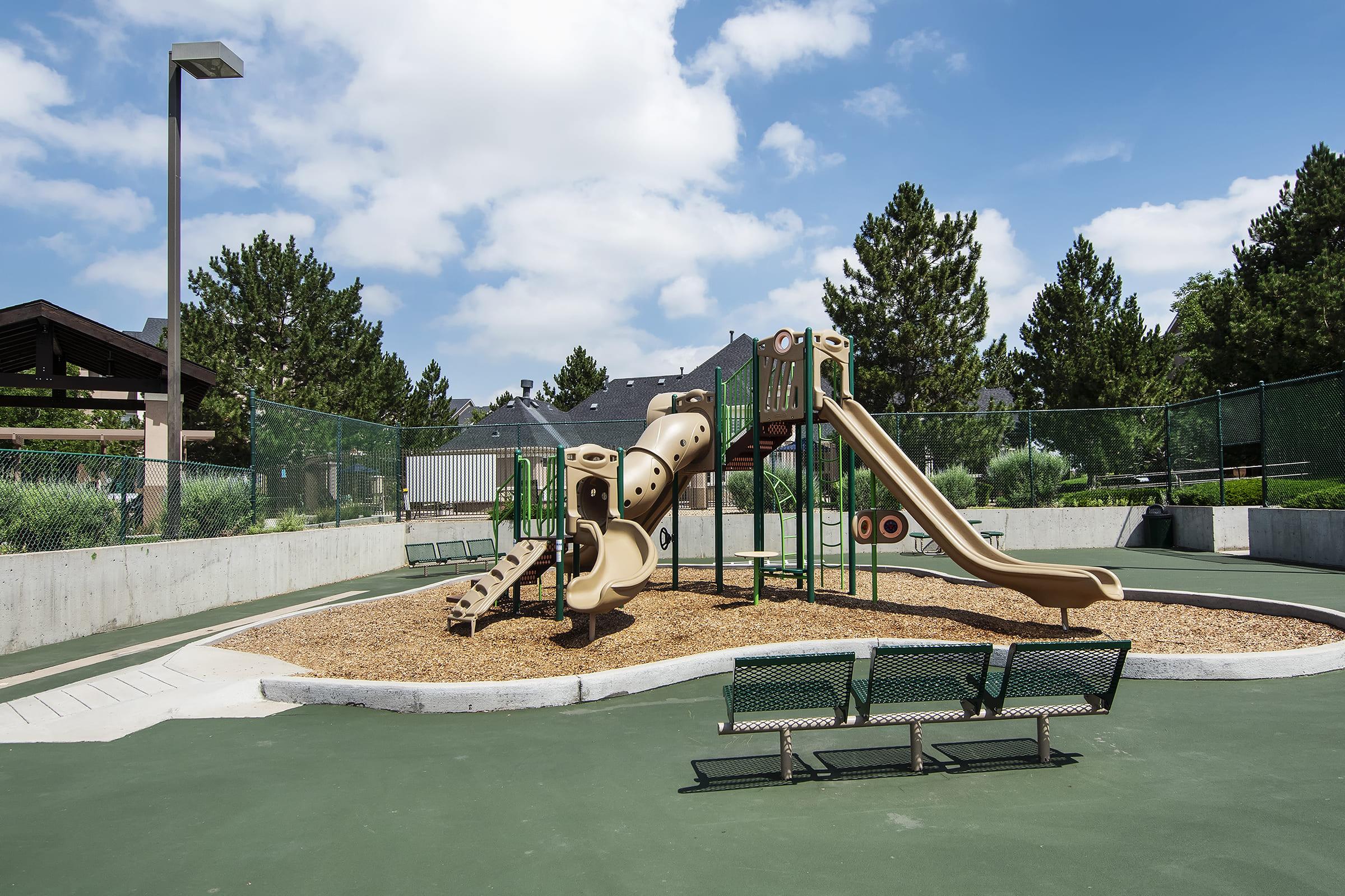 A playground featuring a multi-level slide structure surrounded by tan wood chips. There are two green benches nearby and a fenced area with trees in the background. The sky is partly cloudy, and the playground is set in a well-maintained outdoor space.