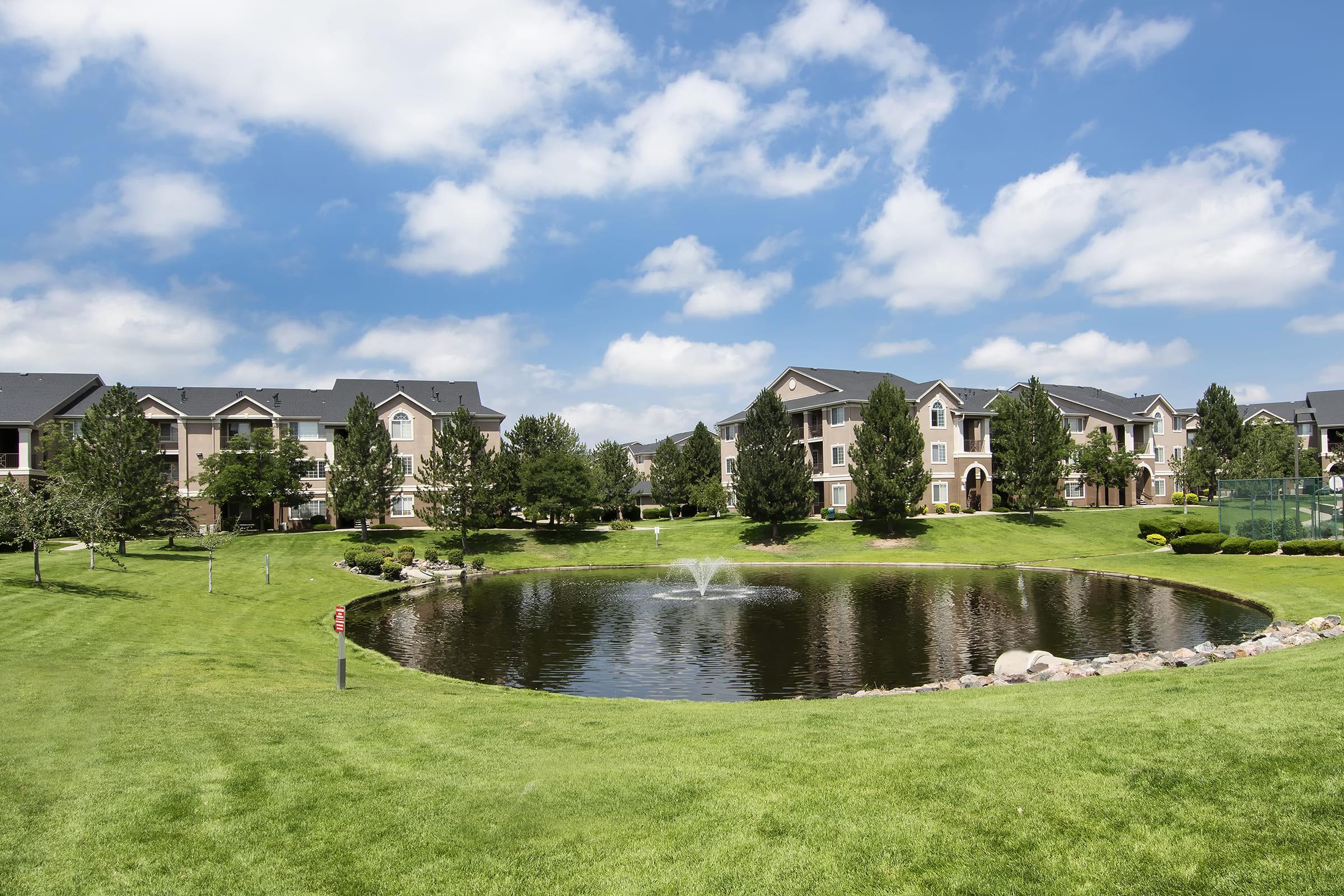 A serene residential area featuring well-maintained apartment buildings surrounded by lush greenery. In the foreground, a tranquil pond with a fountain is bordered by manicured lawns and landscaped trees, under a bright blue sky with fluffy clouds.