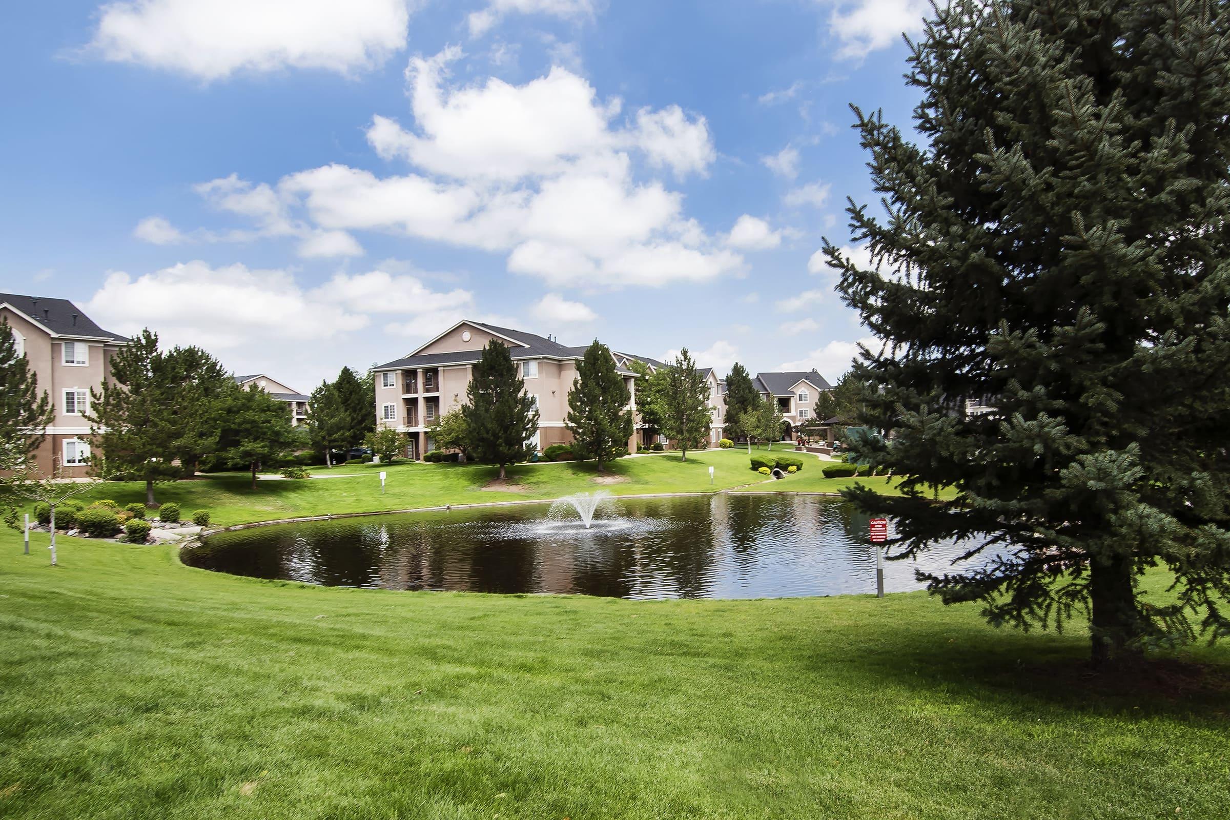 A peaceful residential area featuring well-maintained green lawns, a small pond with a fountain, and several trees. In the background, there are apartment buildings under a blue sky with scattered clouds, creating a serene outdoor environment.