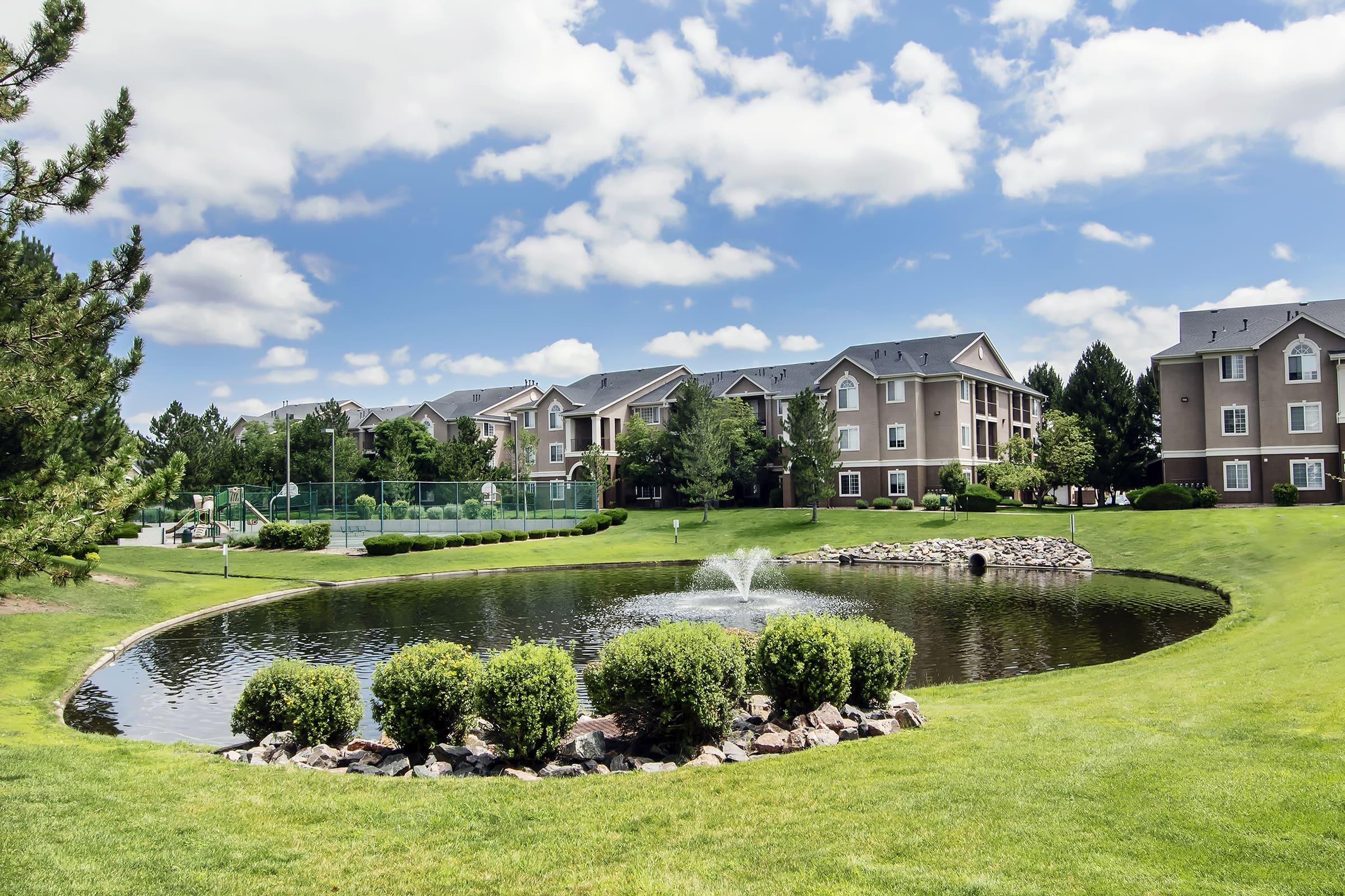 A scenic view of a landscaped area featuring a pond with a fountain, surrounded by grass and shrubs. In the background, there are several multi-story apartment buildings and a tennis court. The sky is partly cloudy, creating a bright and inviting atmosphere.