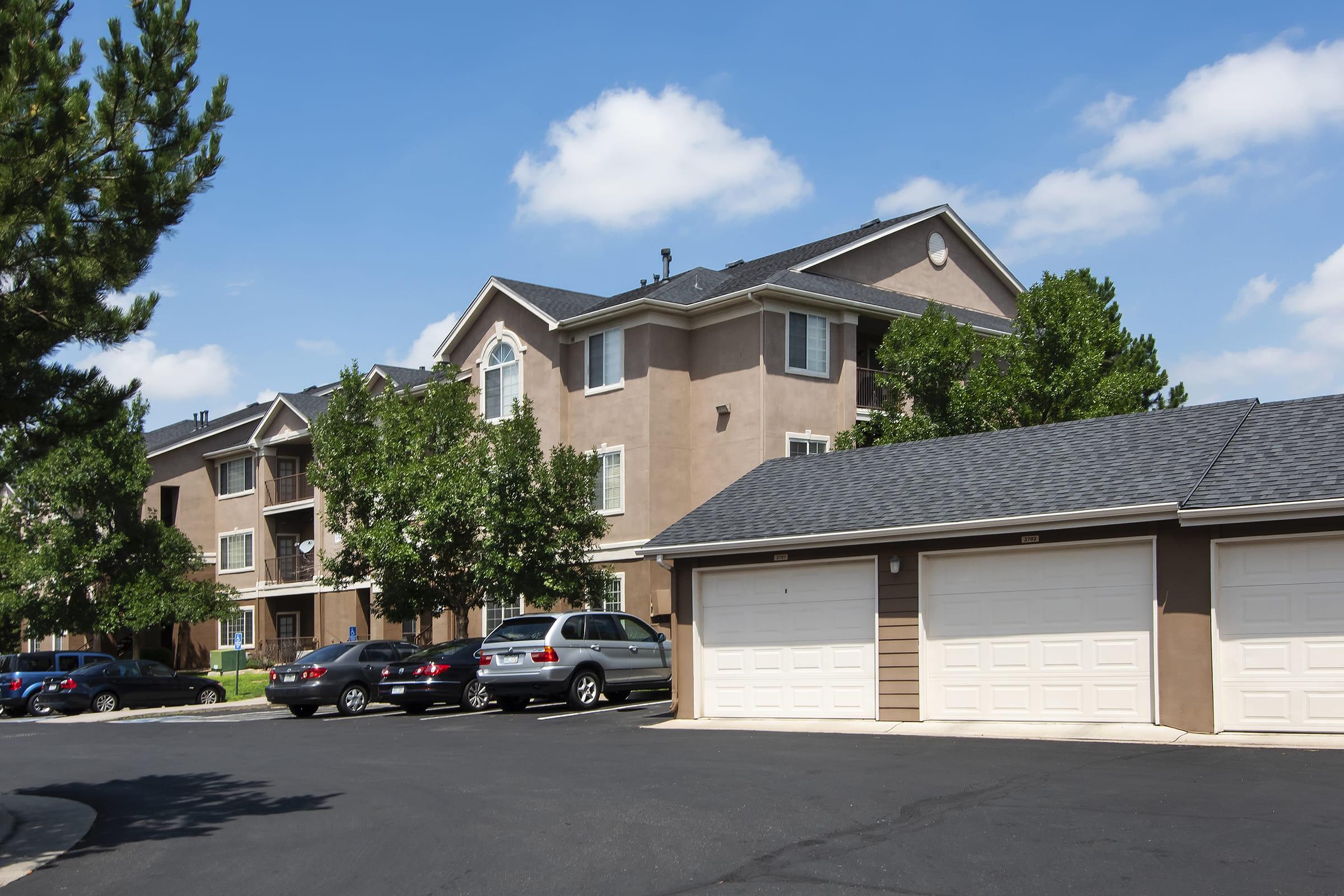 A multi-story residential building with a light brown exterior, surrounded by trees. The foreground features a parking area with several cars parked alongside garages with white doors. The sky is blue with scattered clouds.