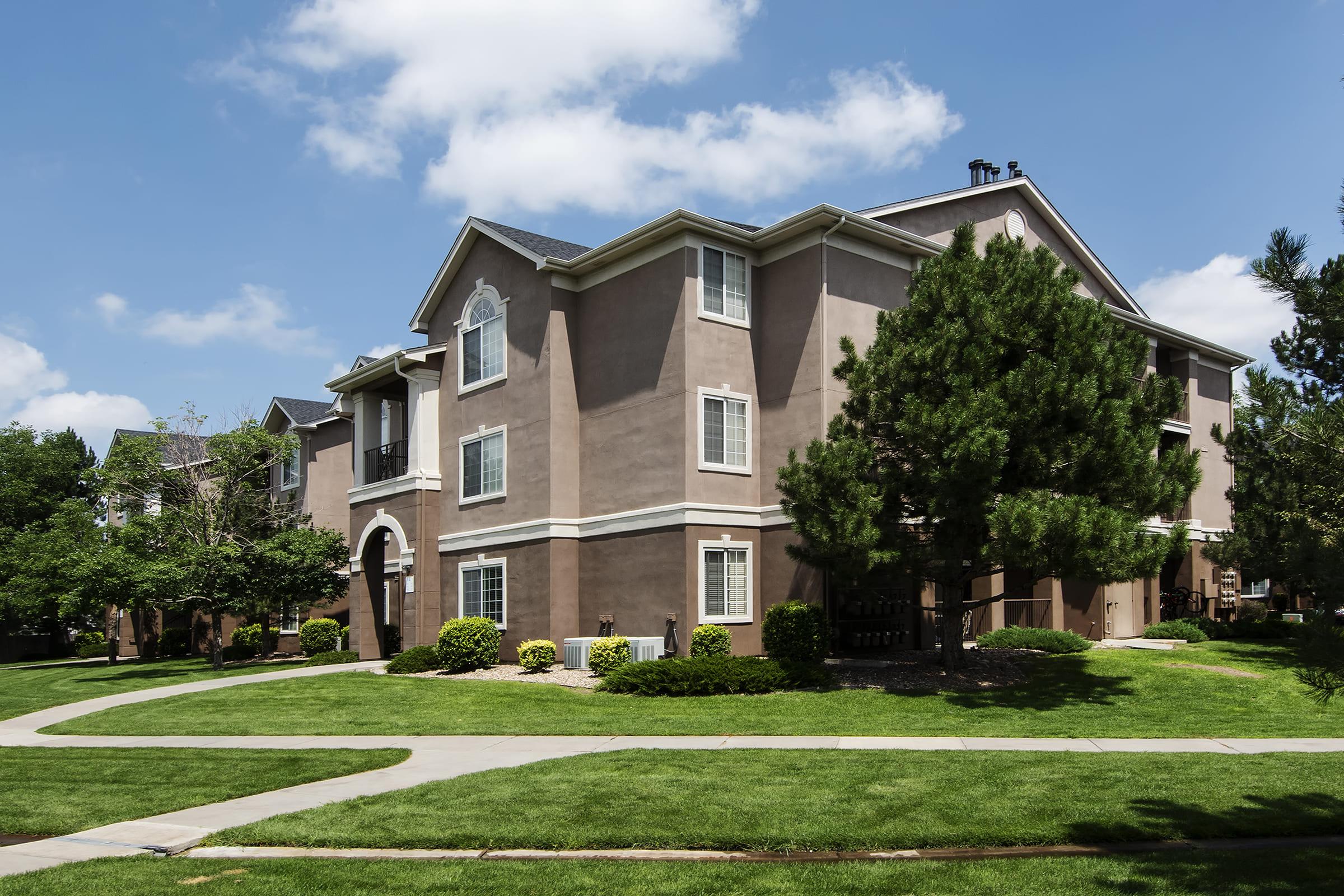 A modern, multi-story apartment building with a light brown exterior, surrounded by well-maintained green lawns and trees under a blue sky with fluffy clouds. A paved path meanders through the landscape, leading to the building's entrance.