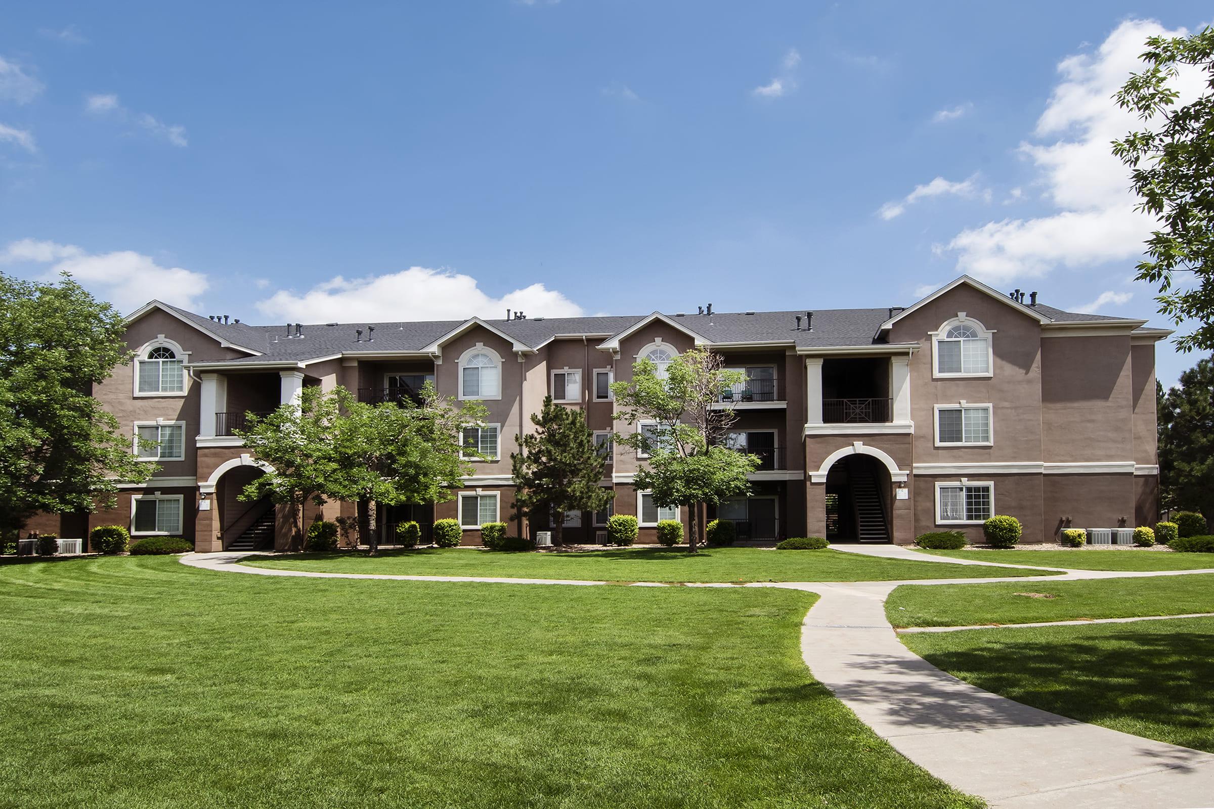 A modern apartment building with multiple levels, surrounded by well-maintained lawns and trees. A paved pathway winds through the green grass, leading to the entrances of the apartments. The sky is clear with a few clouds, indicating a pleasant day.