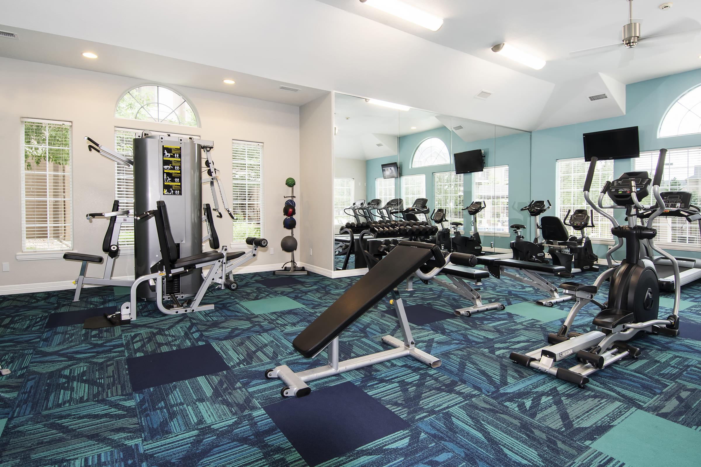 Fitness center featuring a variety of exercise equipment, including a weight machine, a bench for weights, and several cardio machines such as treadmills and ellipticals. The space is well-lit with large windows and mirrors, and has a colorful carpet design.