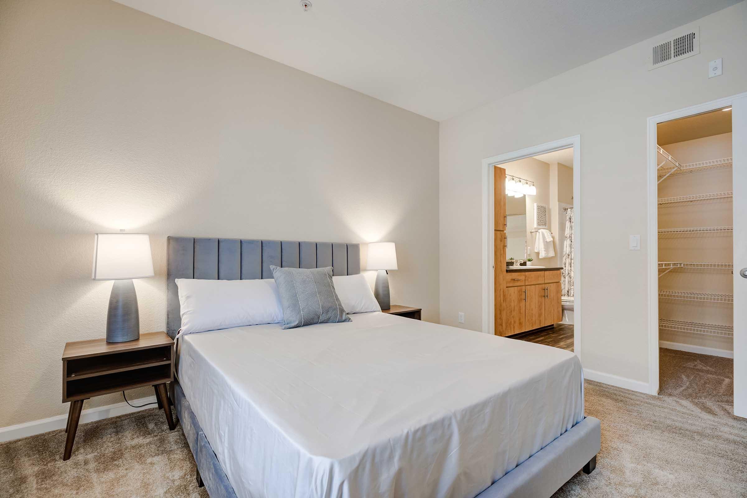 A cozy bedroom featuring a queen-sized bed with gray bedding, matching nightstands with lamps on either side, and a light-colored carpet. A doorway leads to an adjacent bathroom with wooden cabinetry and a well-organized closet, enhancing the room's modern and inviting ambiance.