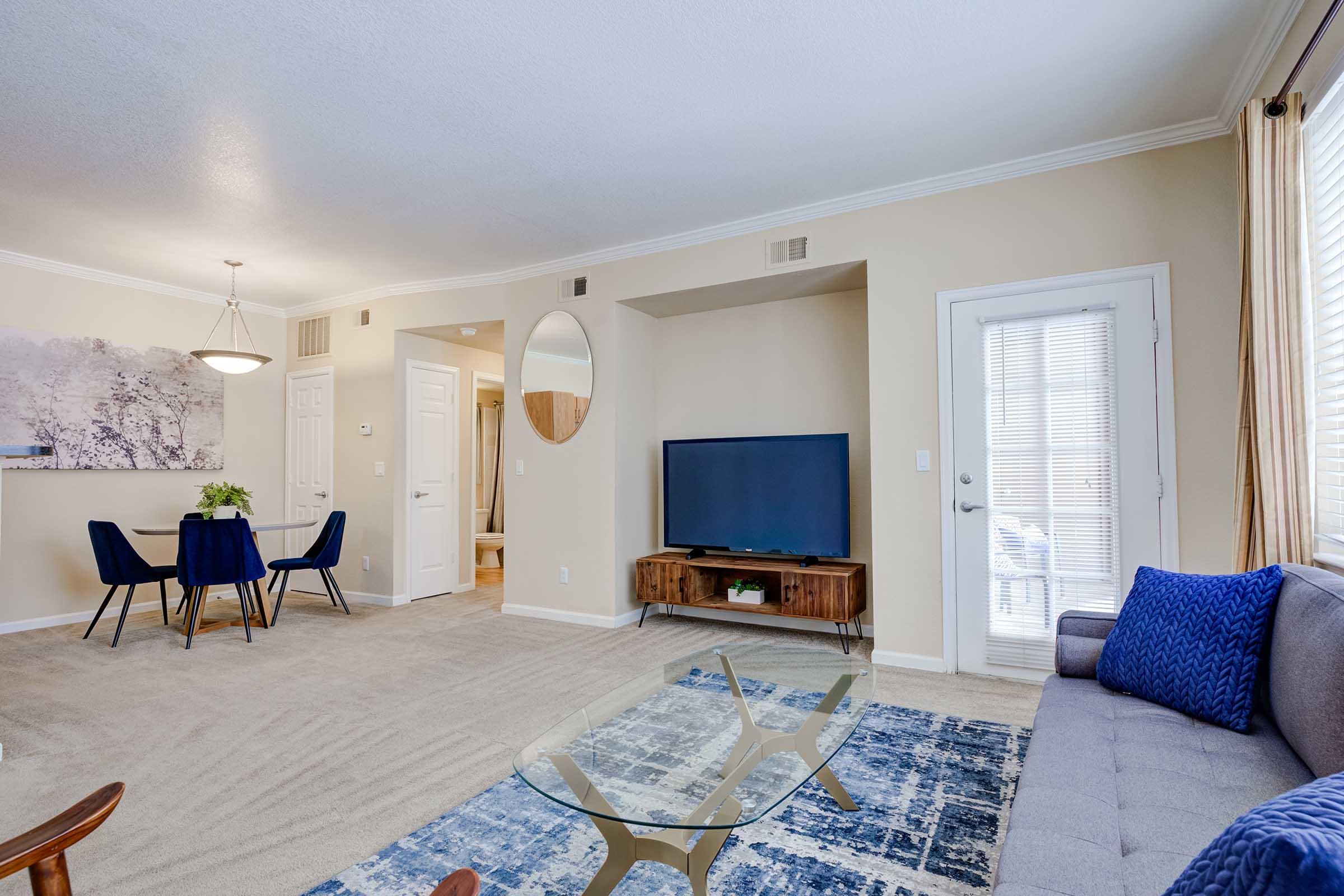 A modern living room featuring a gray sofa with blue throw pillows, a glass coffee table, and a large television on a wooden stand. To the side, there's a dining area with a round table and four chairs. Soft lighting enhances the warm atmosphere, complemented by neutral wall colors and carpeted flooring.