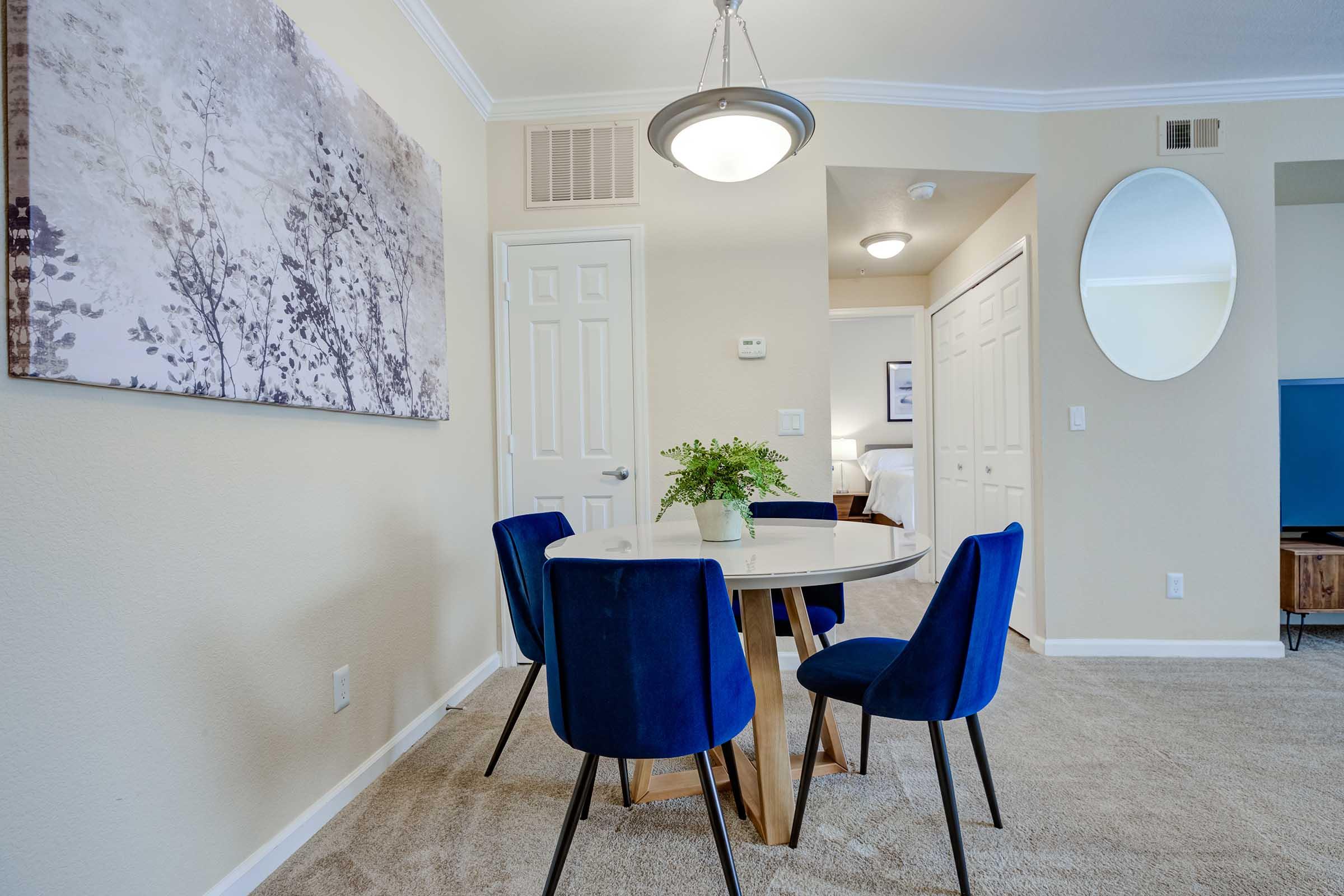 A well-decorated dining area featuring a round wooden table surrounded by four blue upholstered chairs. A floral artwork is hanging on the wall, and there's a small plant centerpiece on the table. The room has beige walls, carpeted flooring, and an elegant light fixture overhead. A rounded mirror and doorway leading to another room are visible.