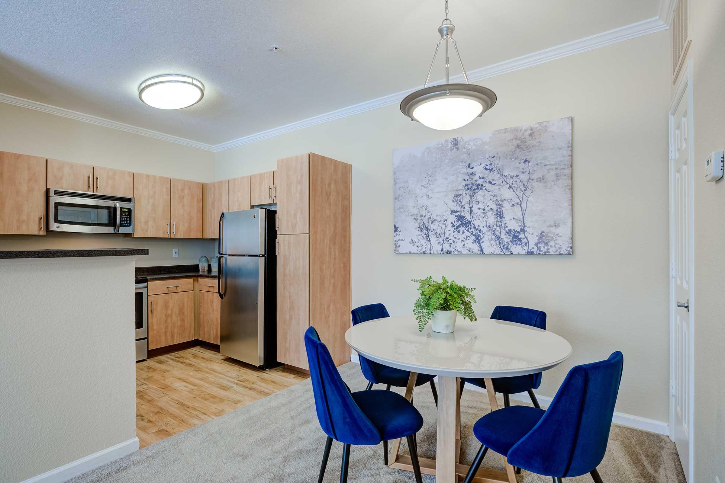 Modern kitchen area featuring light wood cabinetry, stainless steel appliances, and a round white dining table with blue chairs. A large wall art piece complements the neutral wall color. Soft lighting from a ceiling fixture adds warmth to the space.