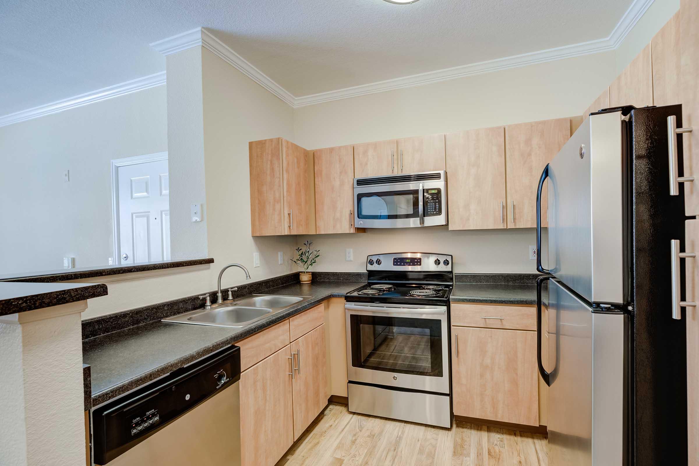 Modern kitchen with light wood cabinetry, stainless steel appliances including a refrigerator, oven, and microwave, and a dark countertop. The room features a double sink and tile flooring, with a spacious layout and neutral wall colors.