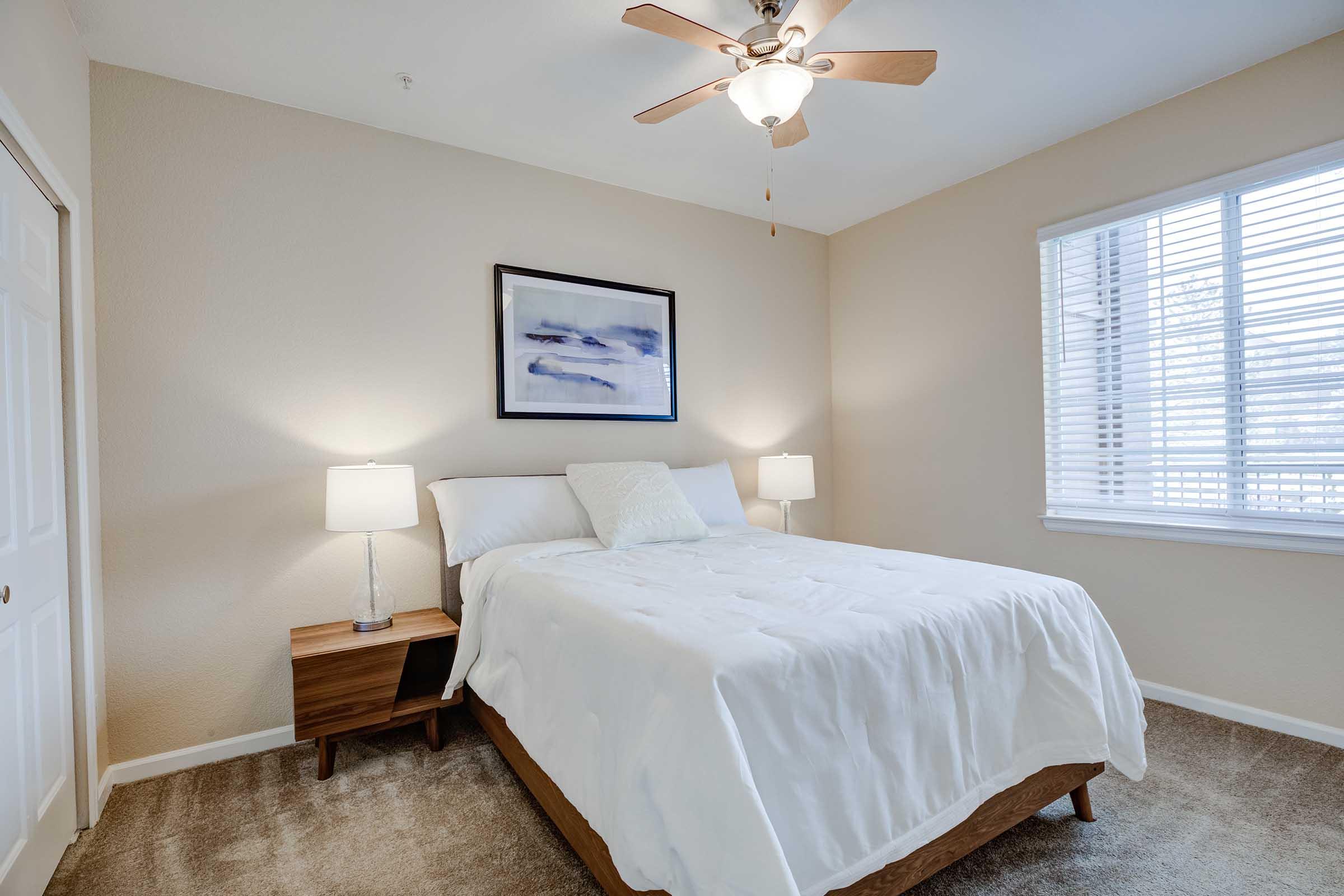 A cozy bedroom featuring a comfortable bed with white bedding, two bedside lamps on wooden nightstands, and a piece of abstract art on the wall. Natural light streams in through a window with blinds, illuminating the warm, neutral-colored walls and soft carpet.