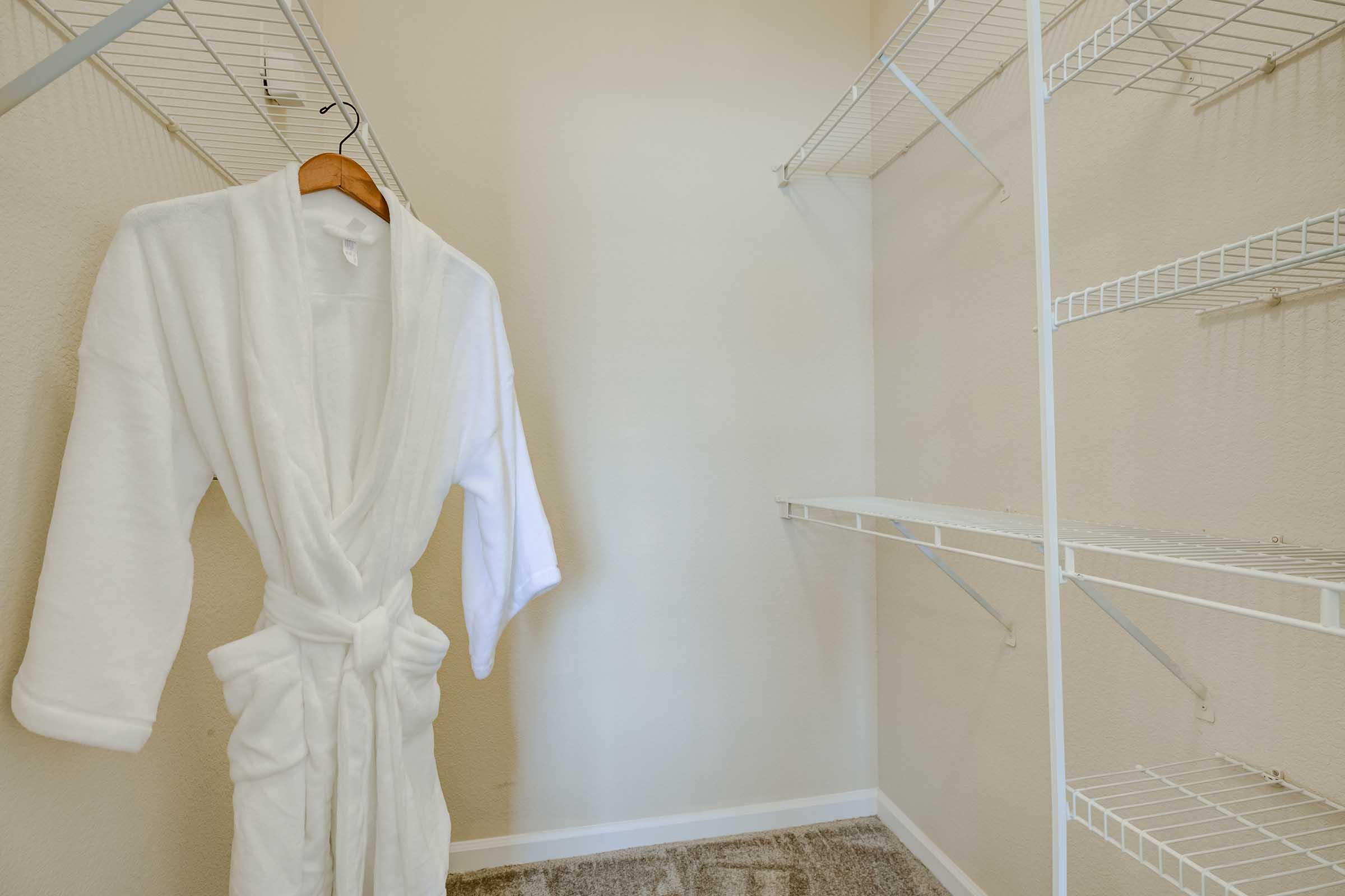A minimalist closet featuring a white bathrobe hanging on a wooden hanger, with empty wire shelves on either side. The walls are painted a soft neutral color, and the floor is carpeted.