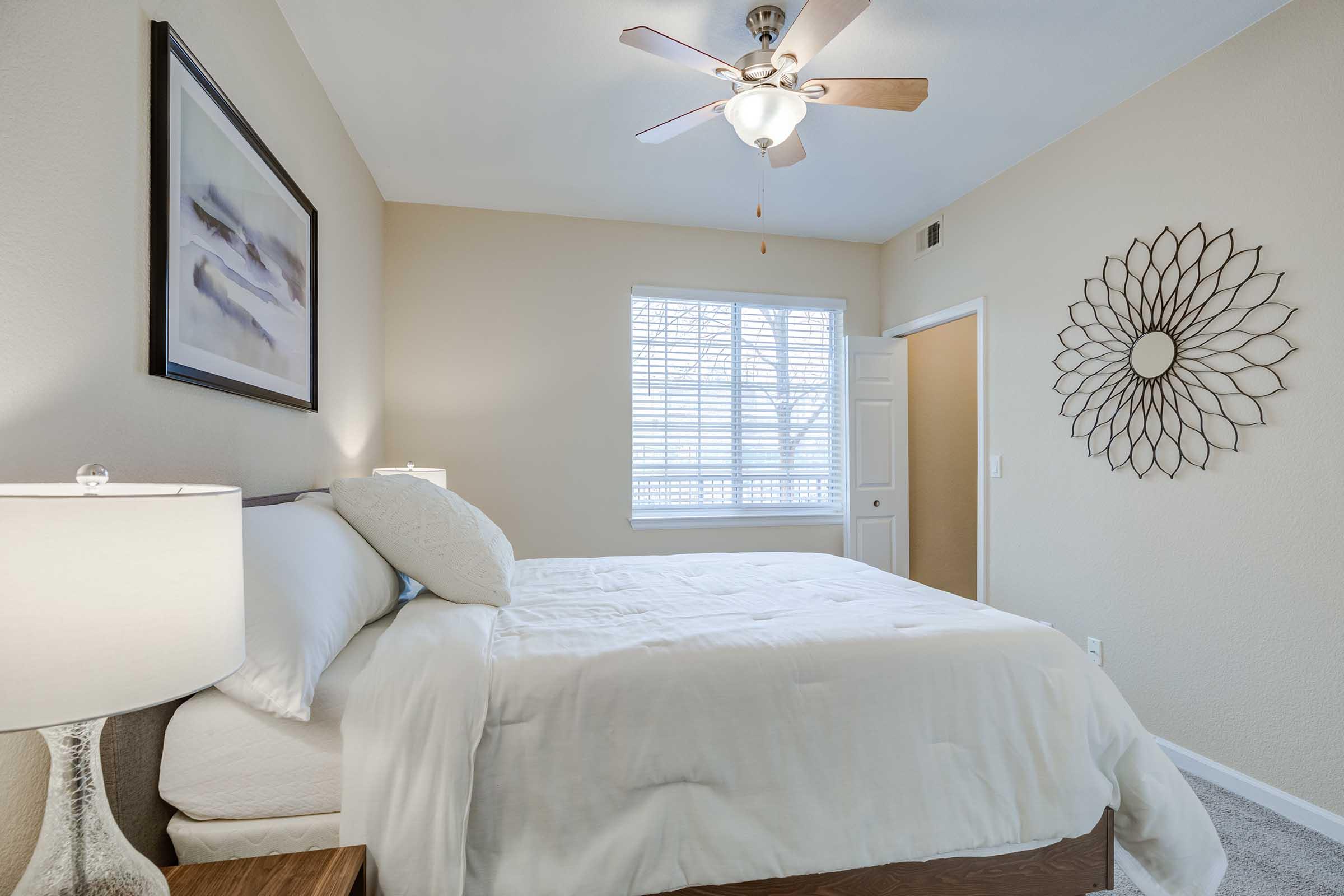 A cozy bedroom featuring a bed with white linens, a decorative lamp on a nightstand, and a large mirror with a floral design on the wall. There's a ceiling fan and a window with blinds allowing natural light to brighten the space, creating a warm and inviting atmosphere.