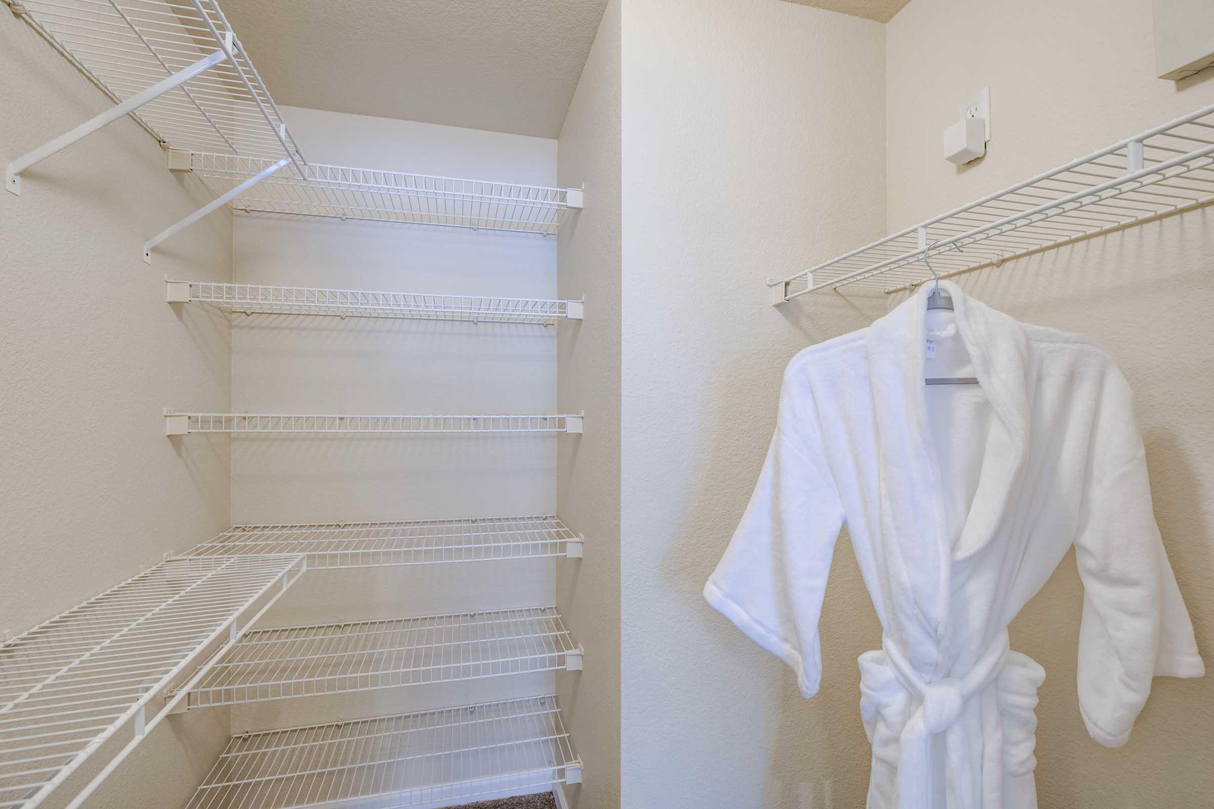 A spacious closet with white wire shelving on the walls, featuring several empty shelves. A white bathrobe hangs on a hanger on one of the shelves, suggesting a cozy and organized space. The closet walls are light-colored, contributing to a clean and airy feel.