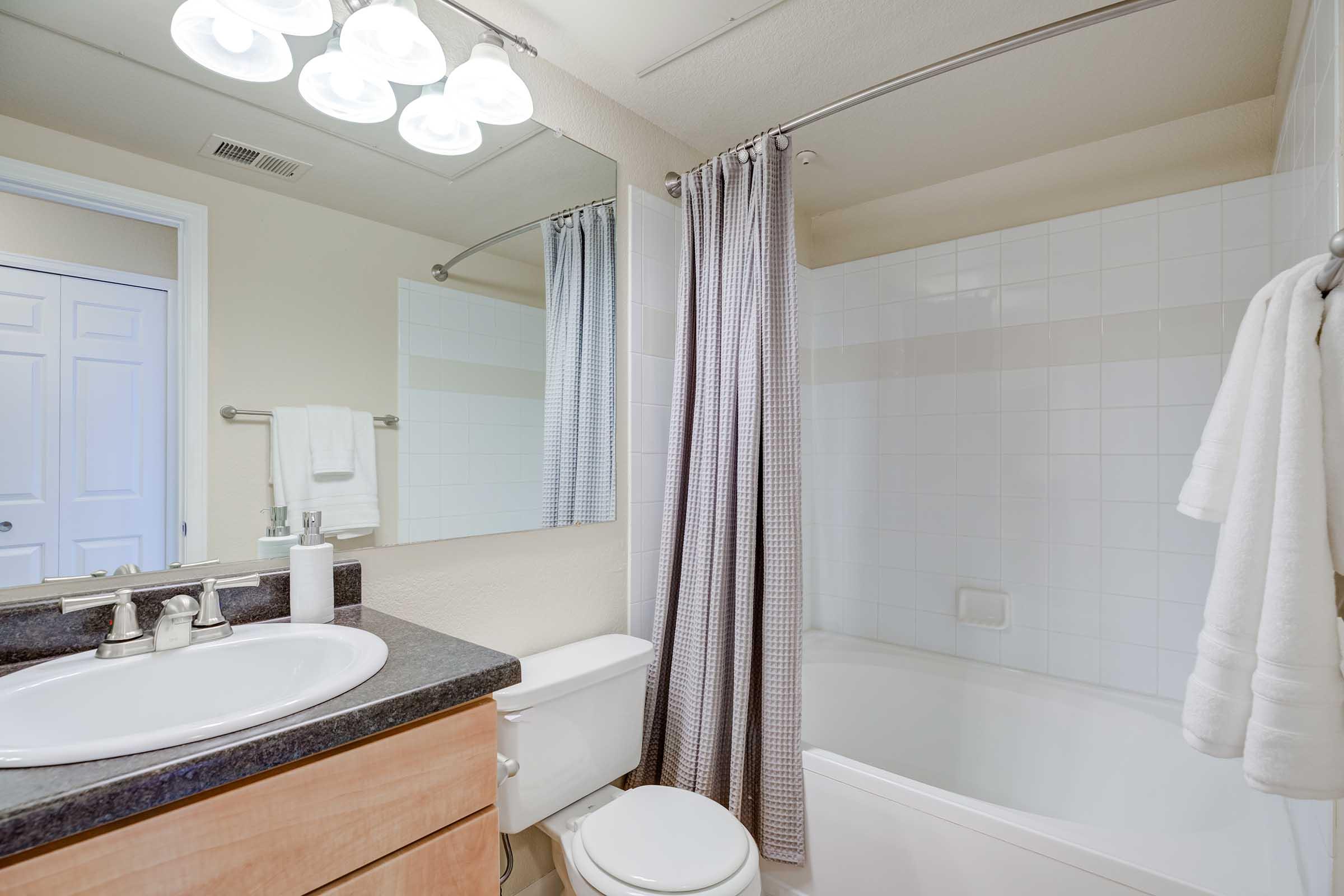 A clean and modern bathroom featuring a white bathtub with a shower, a round sink with a dark countertop, a large mirror above the sink, and a towel rack with white towels. The walls are painted in a neutral color, and a light fixture with multiple bulbs illuminates the space.
