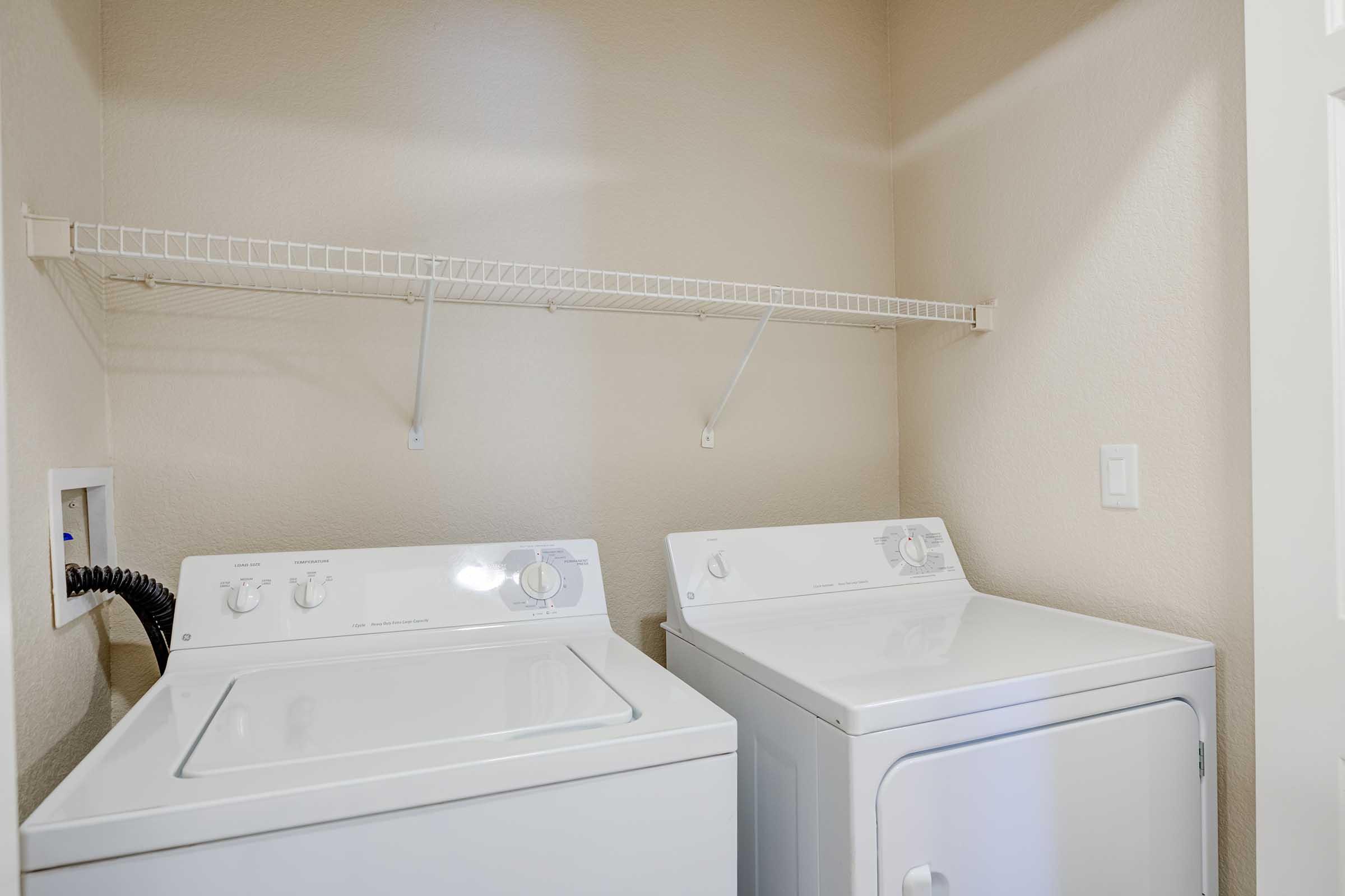 A clean and organized laundry room featuring a washing machine and a dryer side by side. Above them, there's a metal shelving unit mounted on the wall for additional storage. The walls are painted in a light color, creating a bright and spacious feel.