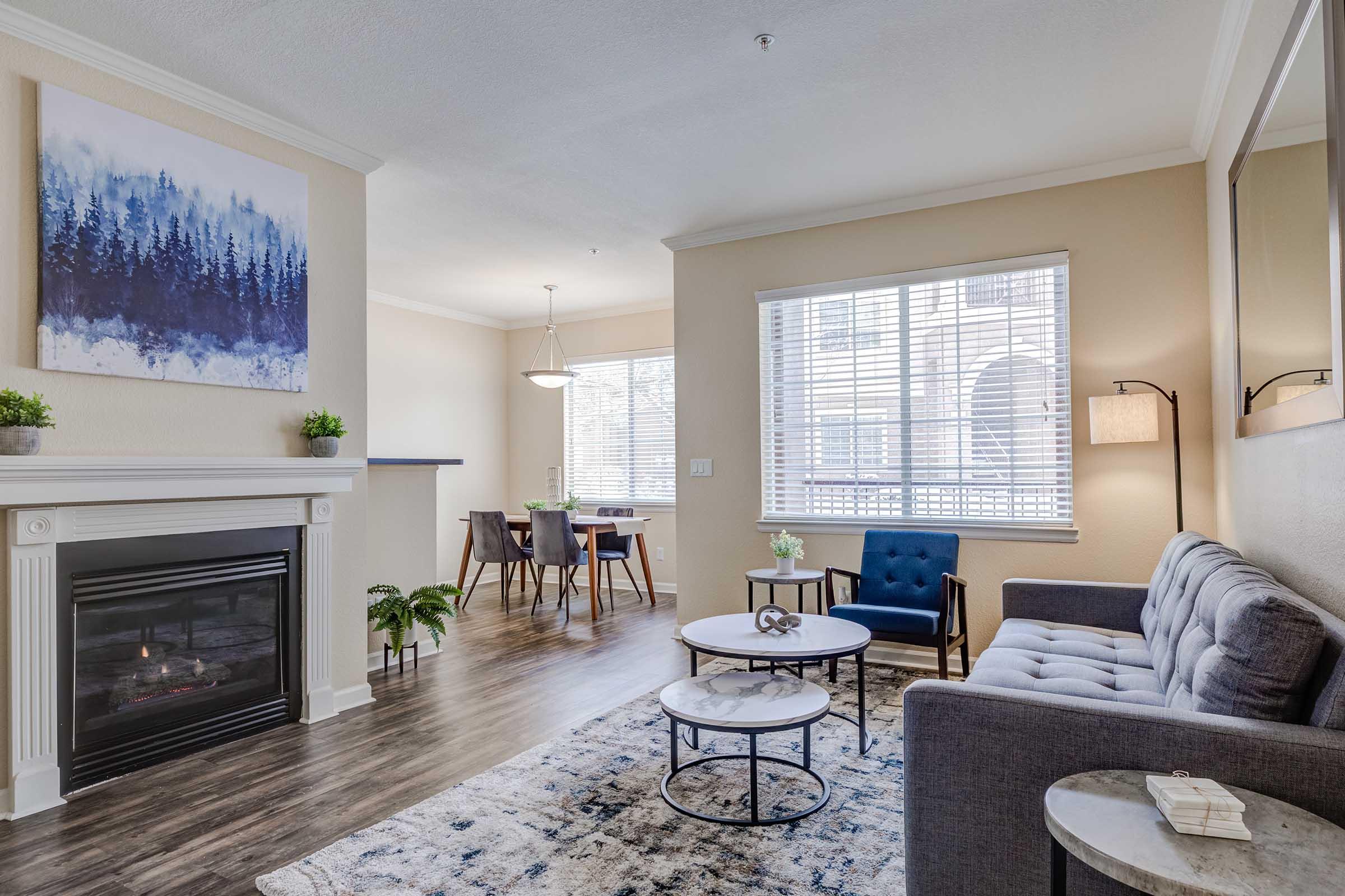 Living room with modern decor featuring a gray sofa, two coffee tables, and a decorative rug. A framed landscape painting hangs on the wall, and there's a fireplace beneath it. Natural light enters through a window, illuminating a dining area with a small table and chairs in the background.