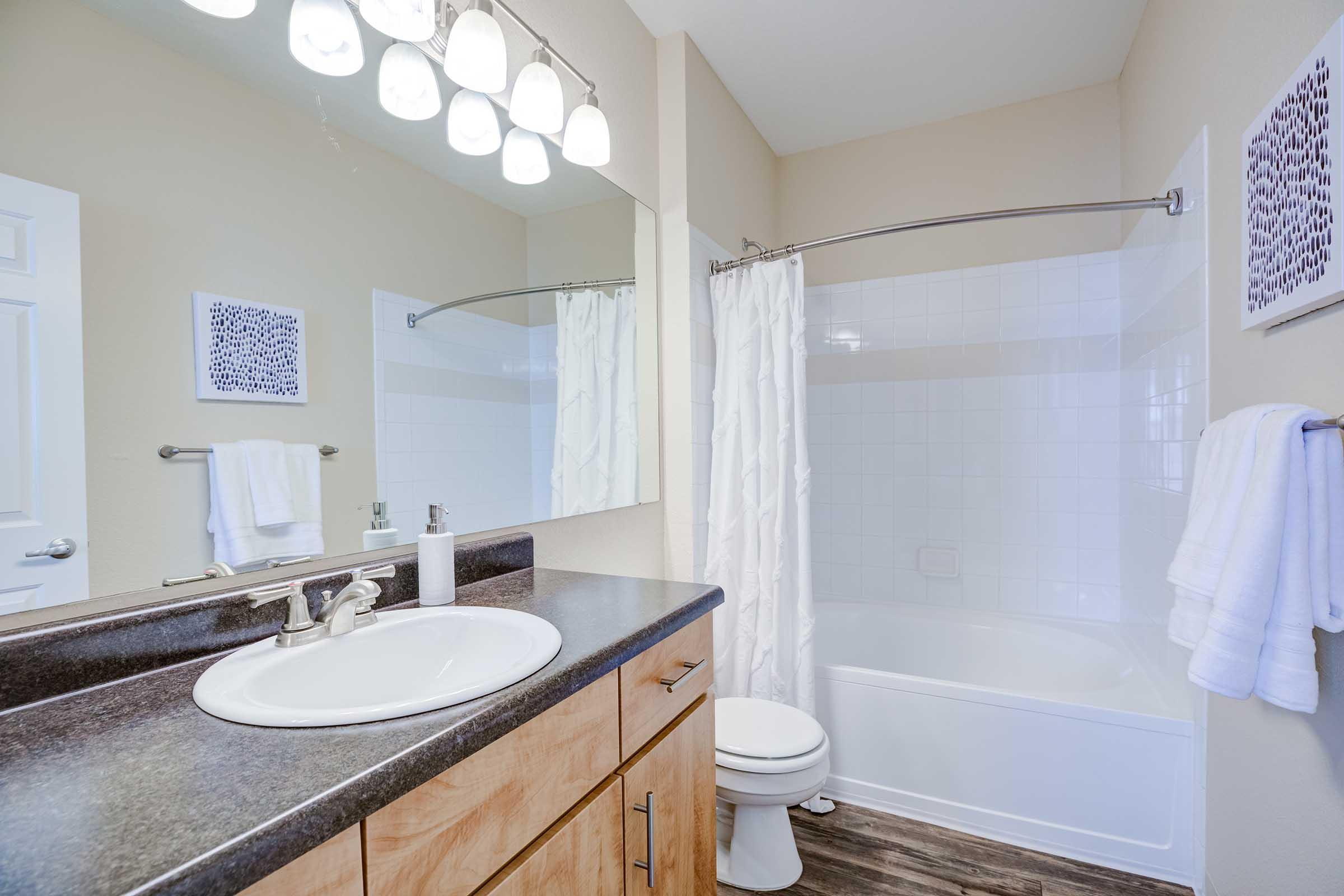 A bright and modern bathroom featuring a white bathtub with a shower curtain, a large mirror with five light fixtures above, a sink with wooden cabinetry, and neatly arranged towels. Soft, neutral colors create a clean and inviting atmosphere.