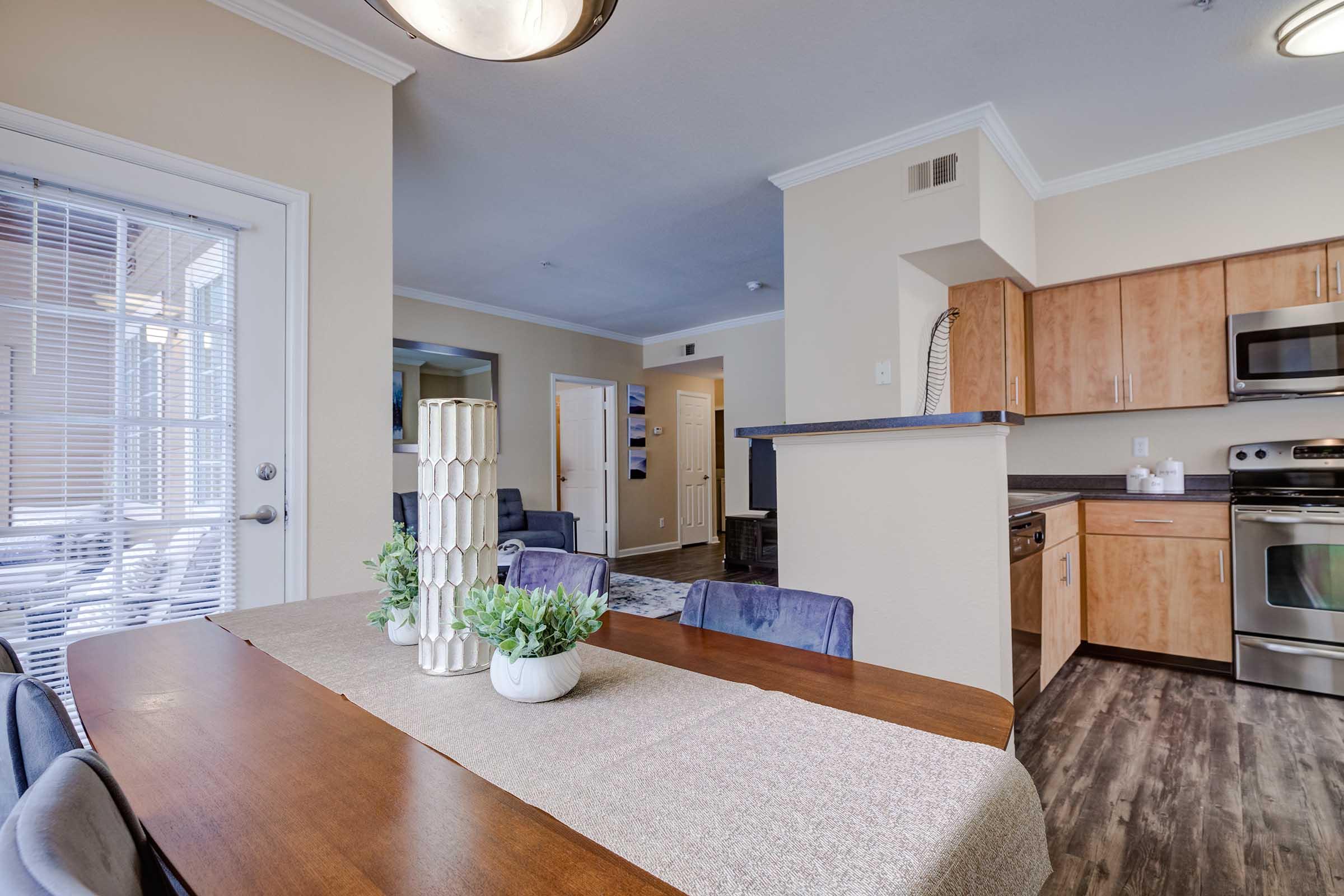 Modern kitchen and dining area with a wooden table set for six, featuring a decorative vase with greenery. Stainless steel appliances in the kitchen, light-colored cabinets, and a large window allowing natural light. A cozy living space is visible in the background, enhancing the open-concept layout.