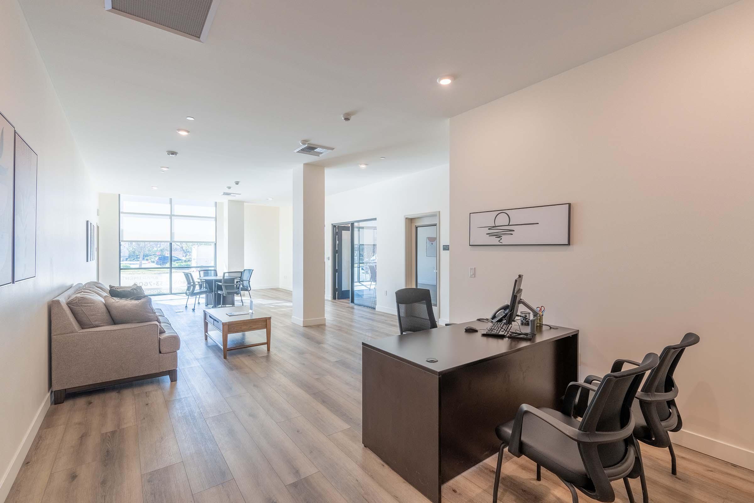 A modern office lobby featuring a reception area with a sleek desk, black chairs, and a comfortable gray sofa. Natural light streams in through large windows, illuminating the light wood flooring. A meeting room is visible in the background with a glass door and a table with chairs.