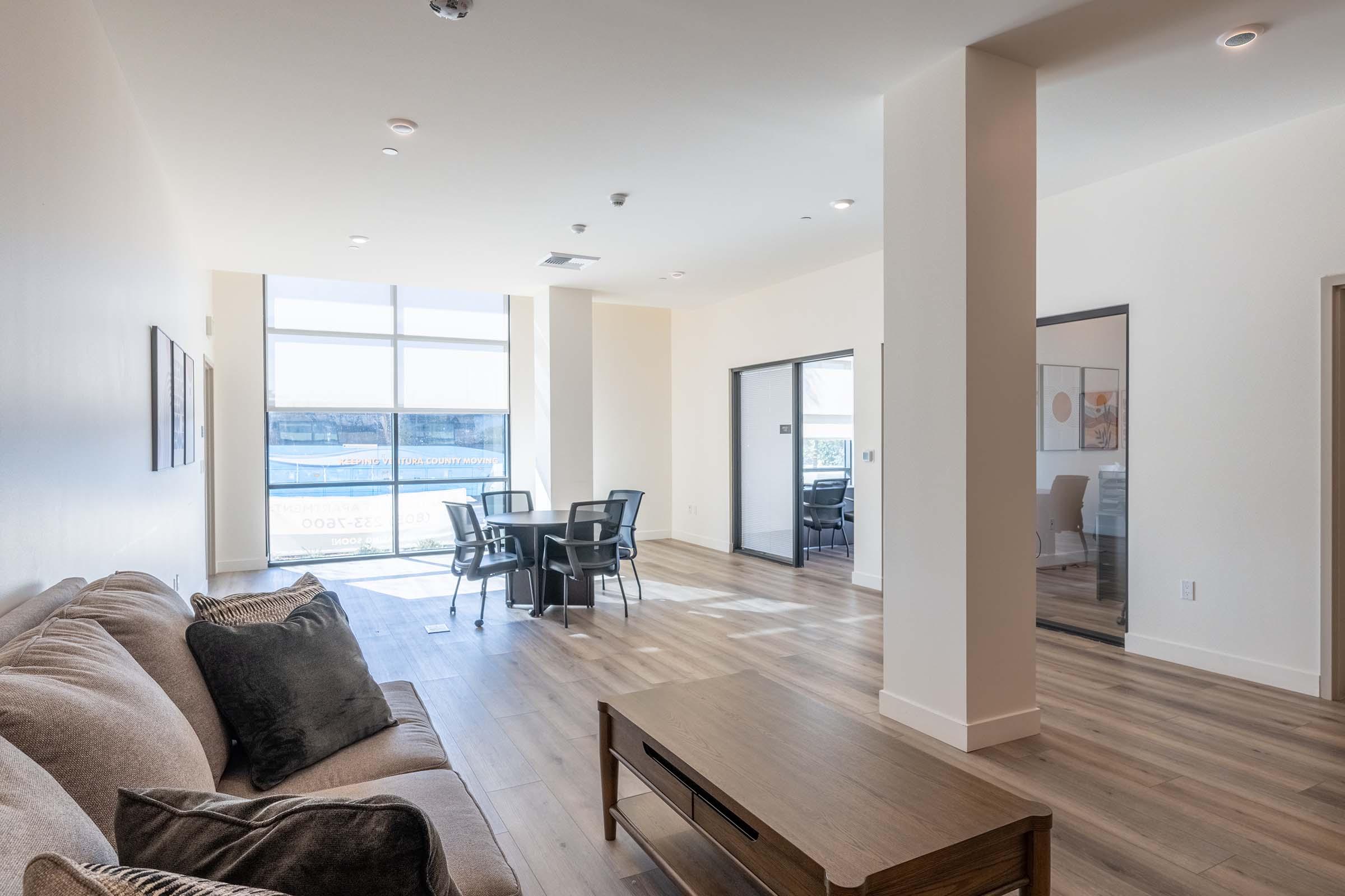 A modern, well-lit living space featuring a cozy sofa with decorative pillows, a wooden coffee table, and a dining area with a round table and four chairs. Large windows allow natural light to fill the room, highlighting the sleek, wooden flooring and minimalist design. A glass door leads to another room.