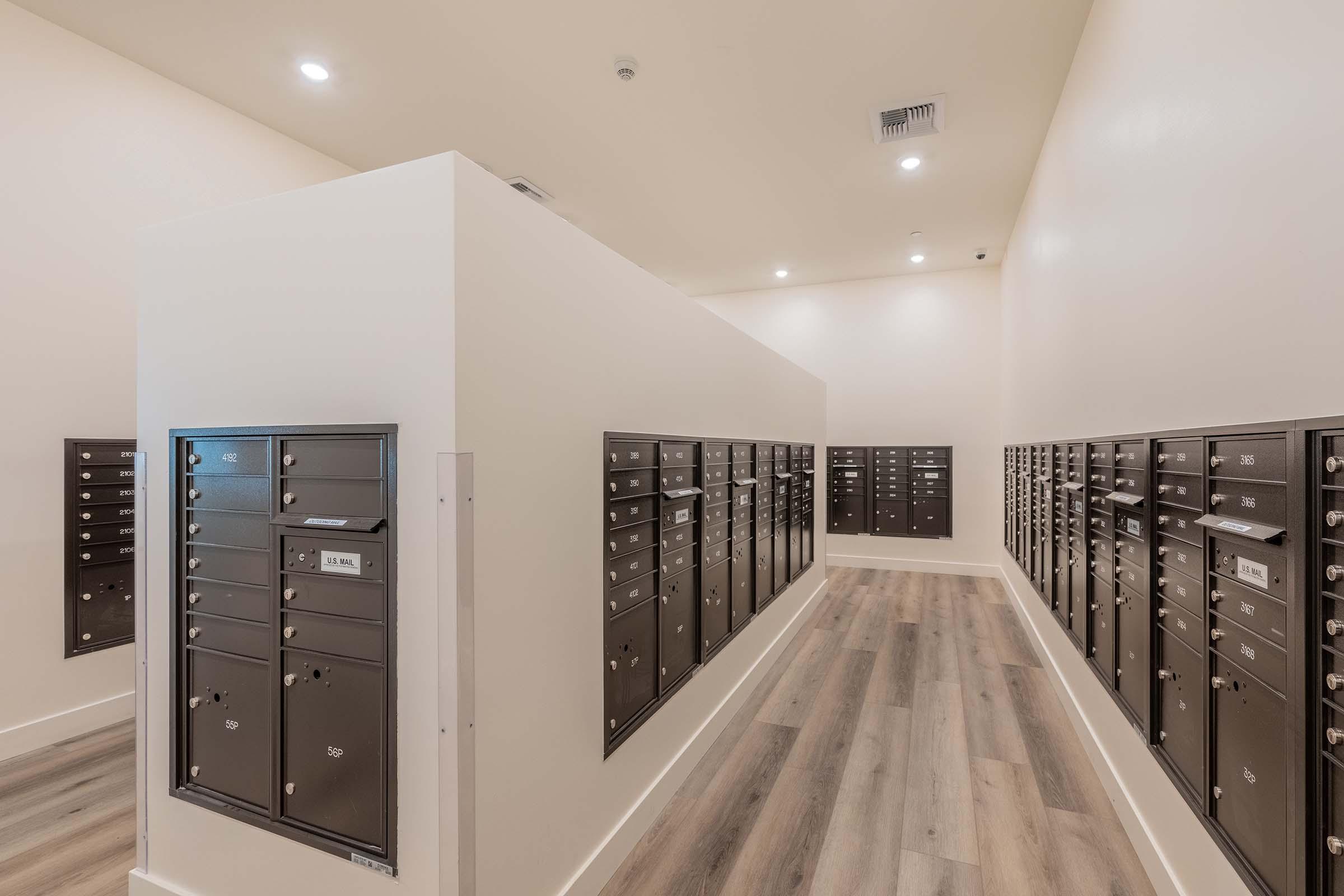 A brightly lit postal room featuring multiple rows of black mailboxes mounted on white walls. The floor has light wooden planks, and the space is designed for efficient mail delivery. The layout is open and modern, providing easy access to the mailboxes.