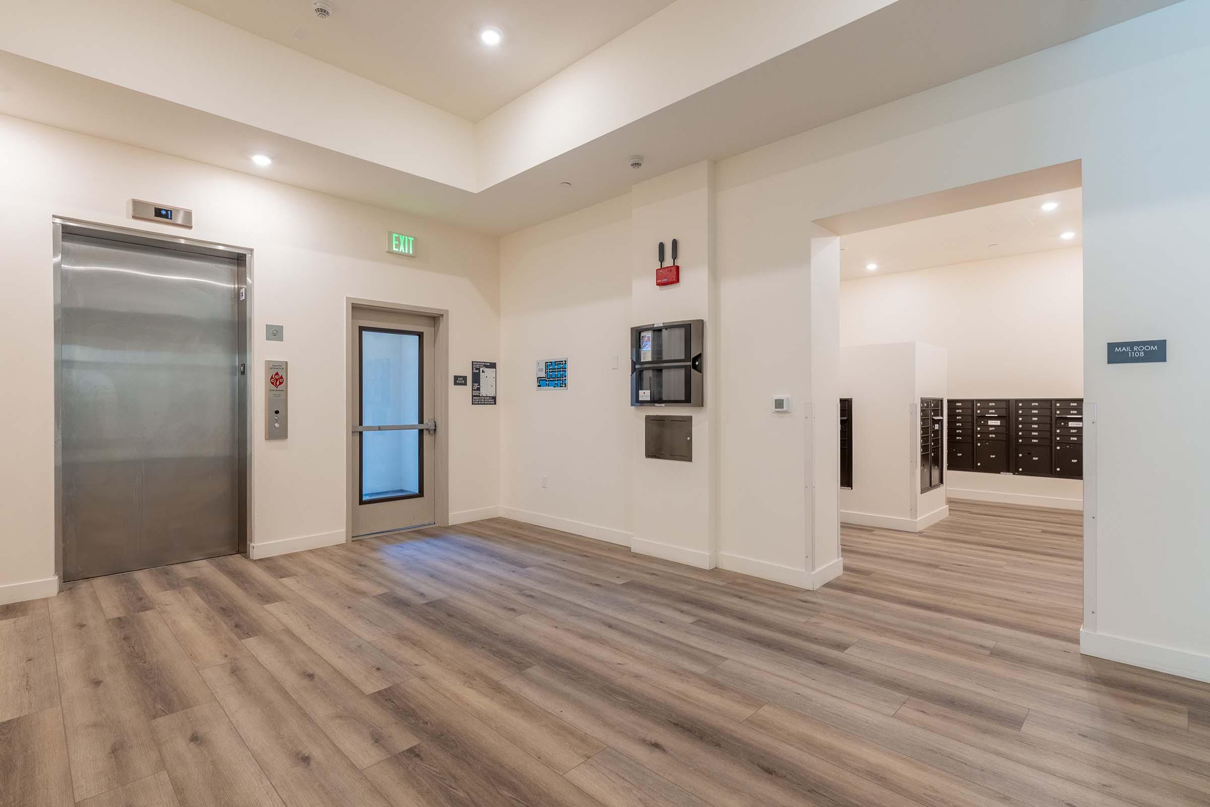 Interior view of a modern hallway featuring an elevator, a door leading outside, and mailboxes. The floors are wooden, and the walls are painted white. Soft lighting creates a welcoming atmosphere.
