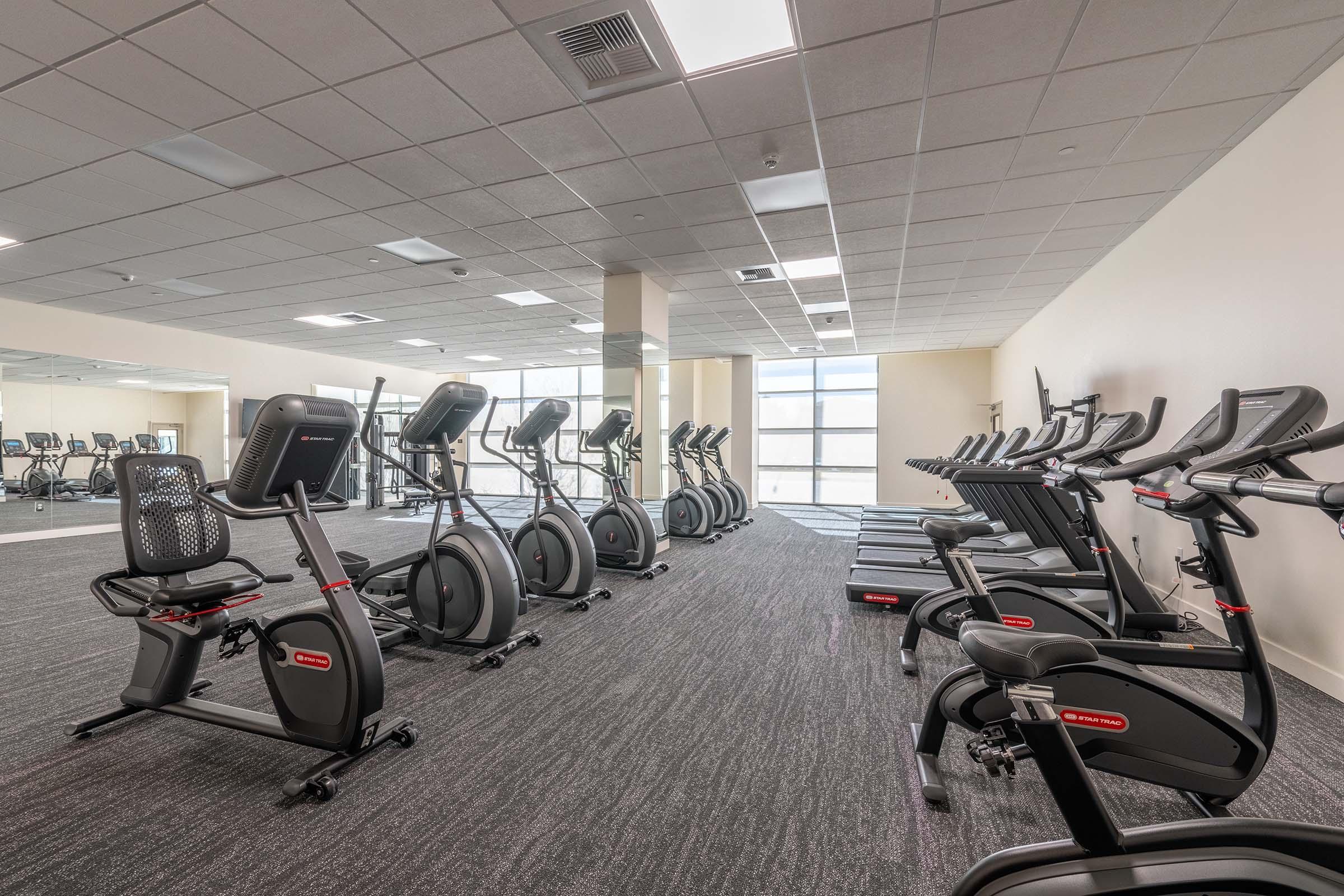 A modern fitness center featuring several exercise machines including stationary bikes, treadmills, and elliptical machines, arranged neatly in a well-lit space with large windows. The flooring is dark and carpeted, creating a sleek and inviting atmosphere for workouts.