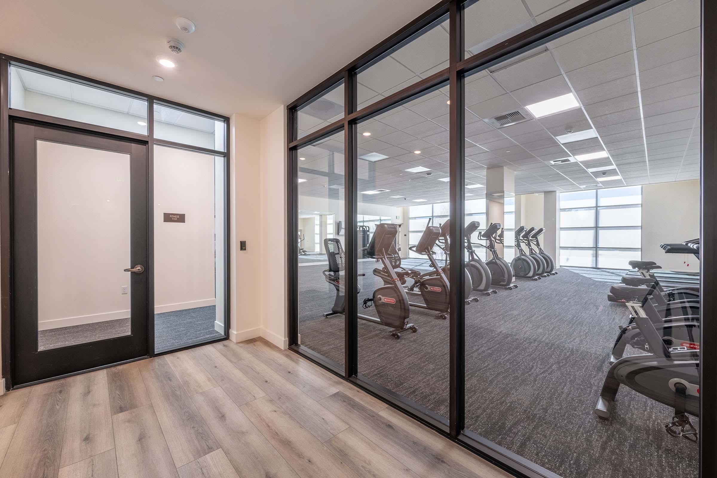 A modern fitness center viewed from a corridor, featuring large glass windows. The gym is equipped with various exercise machines, including treadmills and ellipticals, set against a bright, well-lit space with wooden flooring.