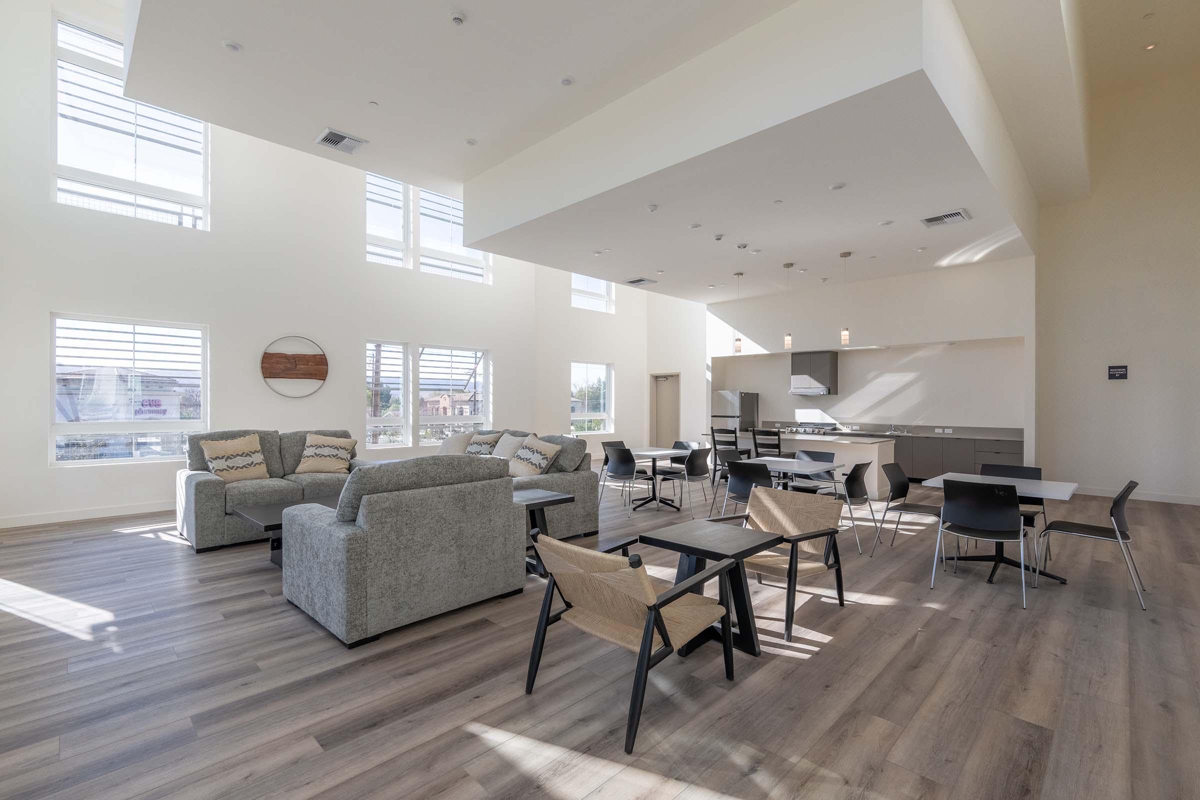 A modern, well-lit communal lounge with high ceilings and large windows. The space features comfortable grey sofas, wooden tables, and black dining chairs. A minimalist kitchen area is visible in the background, creating a welcoming environment for gatherings or relaxation.