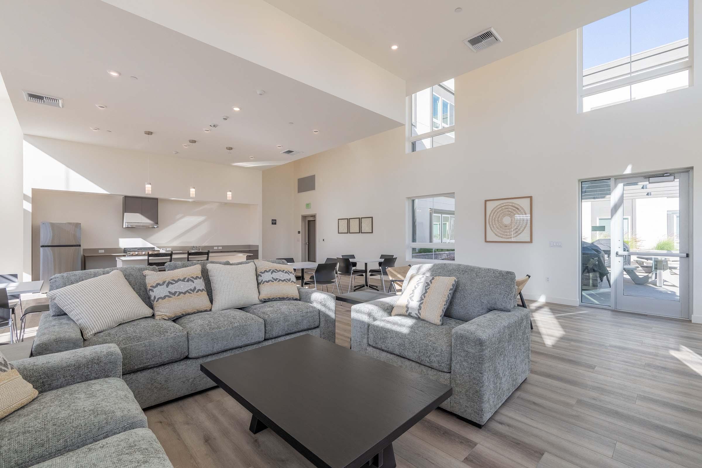 A spacious, modern living room with high ceilings and large windows, featuring a gray sectional sofa with decorative pillows, a dark coffee table, and a dining area in the background. The kitchen is partially visible with stainless steel appliances. Bright natural light floods the space.