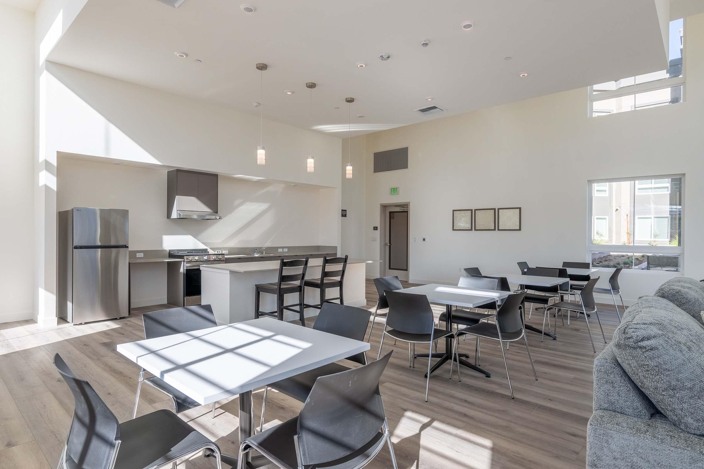 Bright and spacious common area featuring modern furniture. The room includes a kitchen with stainless steel appliances, several tables and chairs arranged for dining or meetings, and large windows allowing natural light to fill the space. A cozy seating area is visible, enhancing the inviting atmosphere.